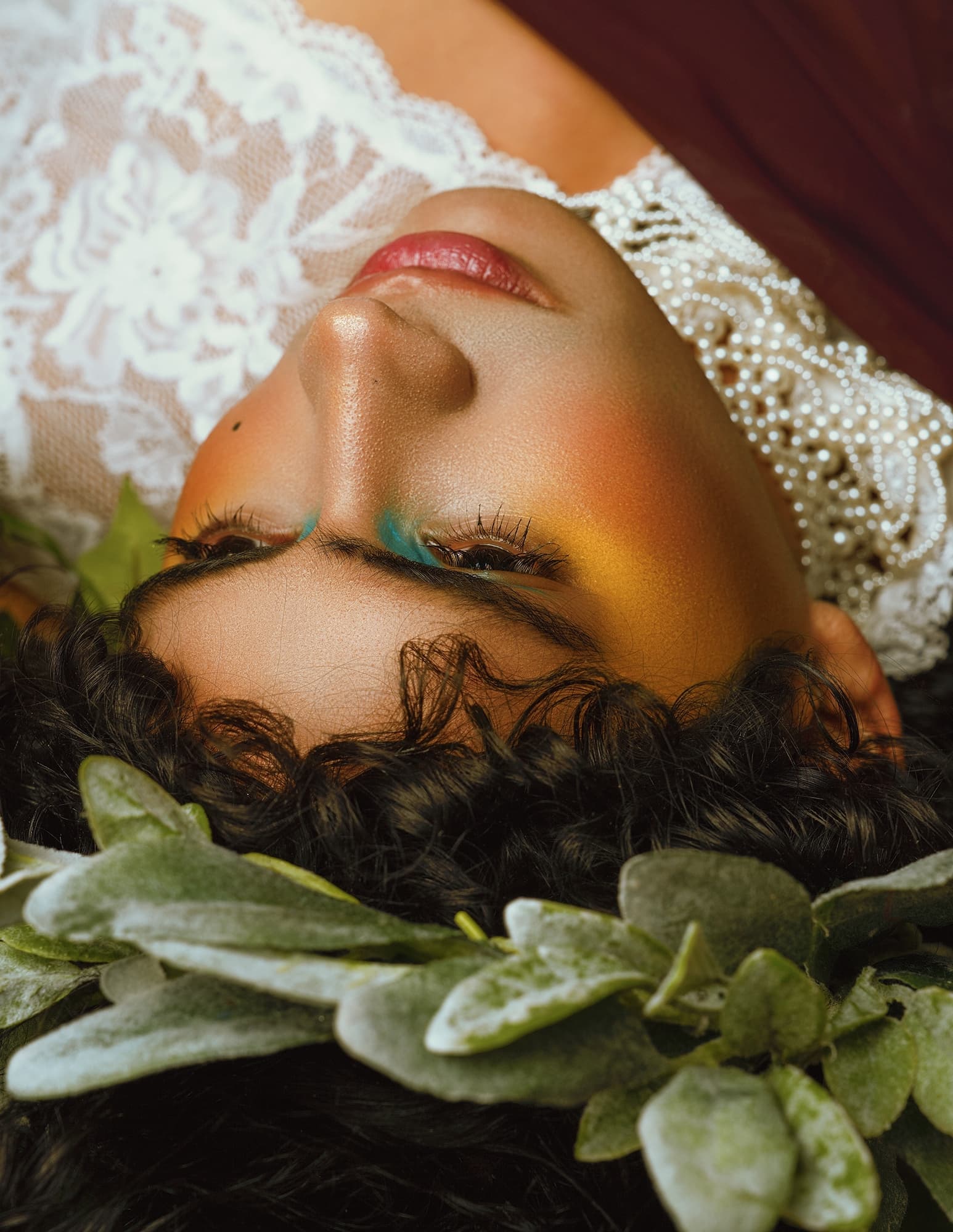 Person with vibrant makeup and crown of leaves, wearing lace, in a serene pose.