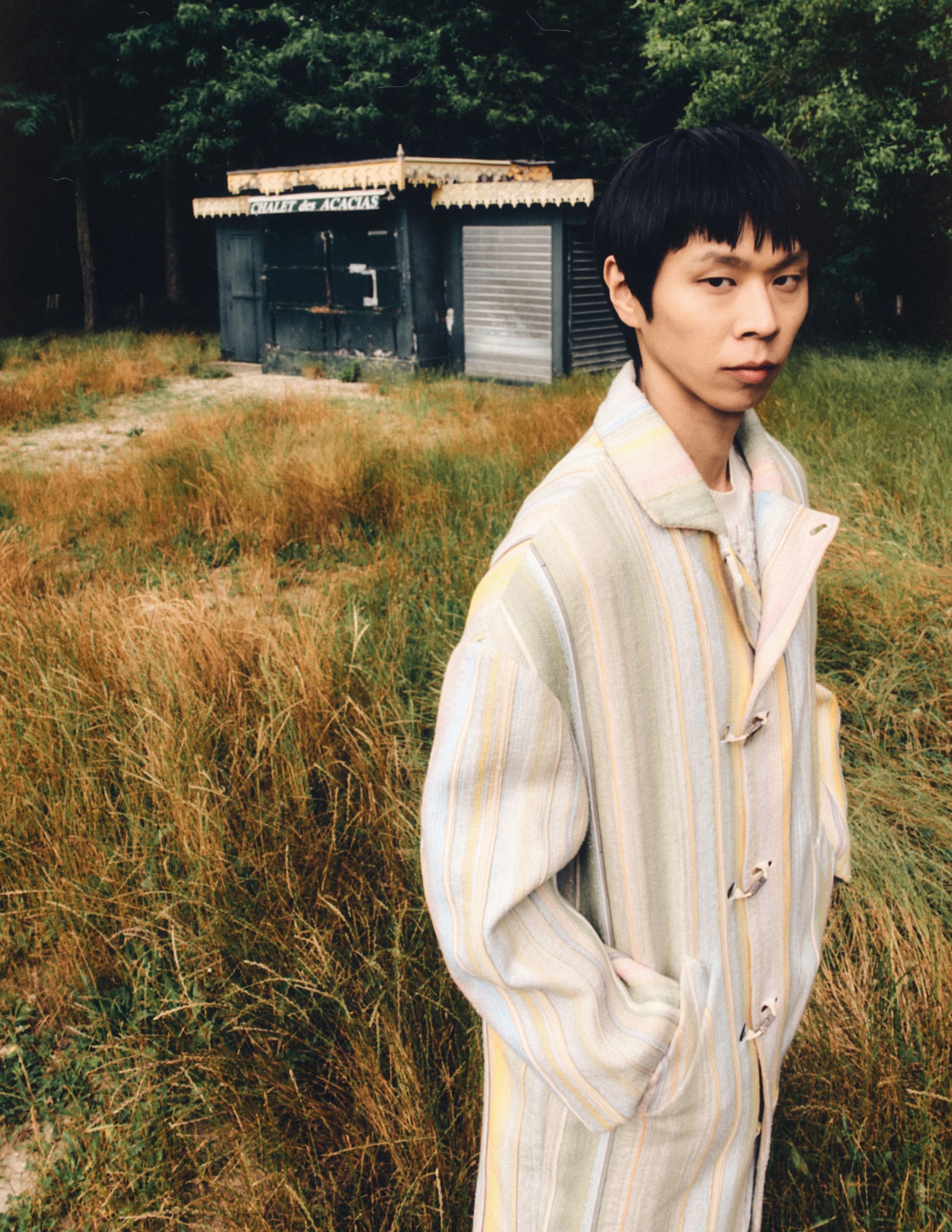Person in striped coat standing in grassy field near rustic kiosk.