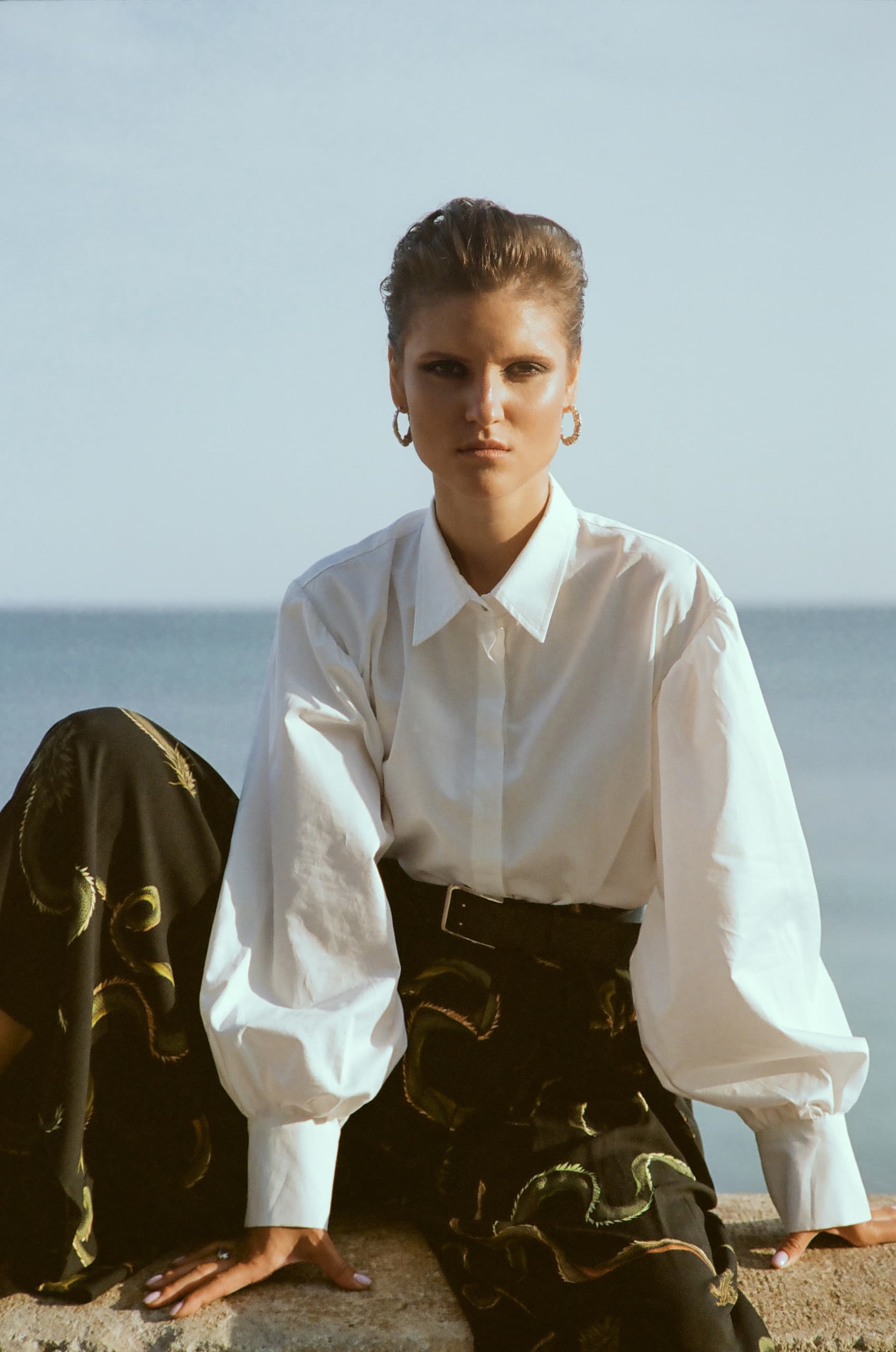 Fashionable woman in white blouse by the sea, wearing hoop earrings.