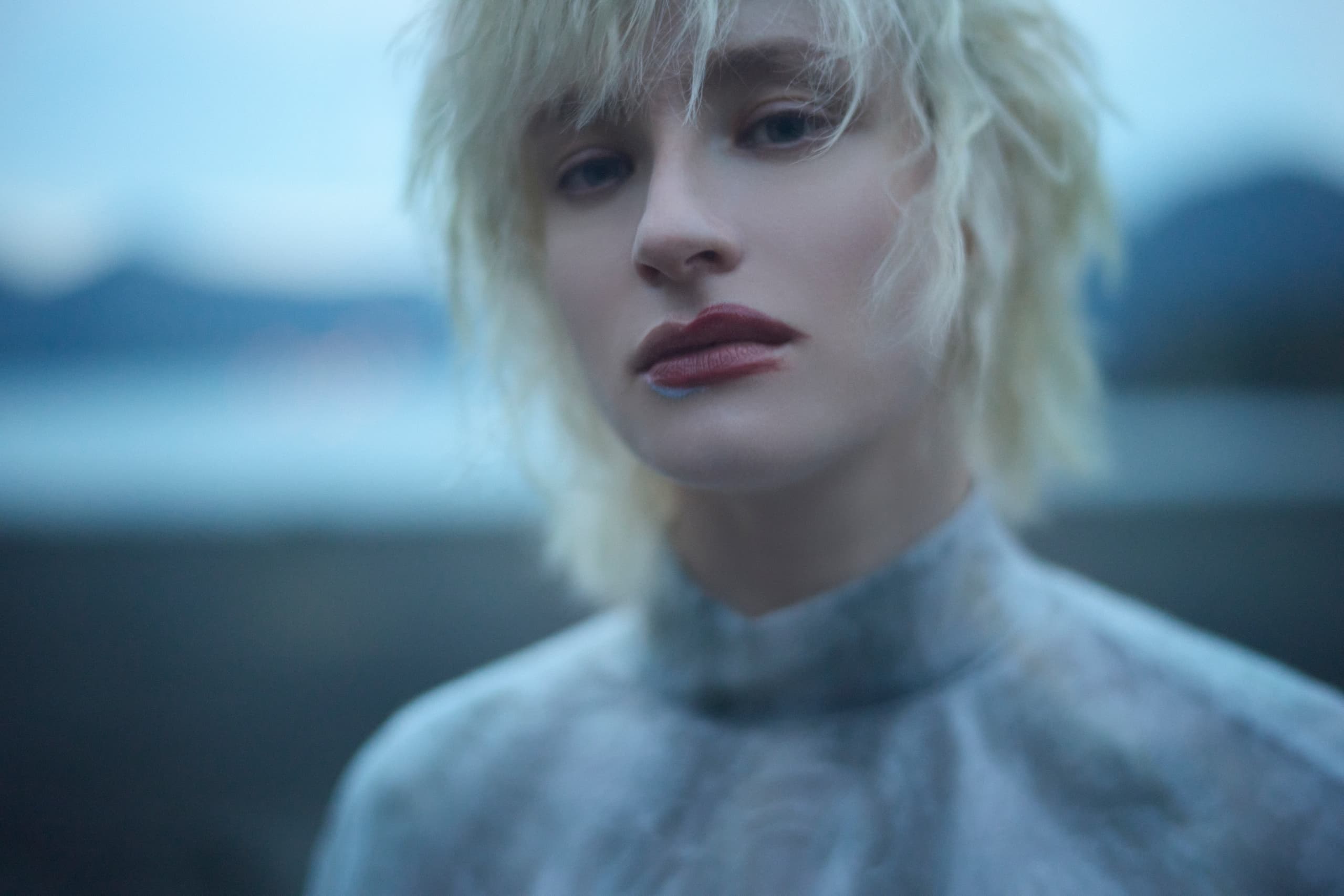 Blonde person with red lipstick in a soft-focus outdoor setting.