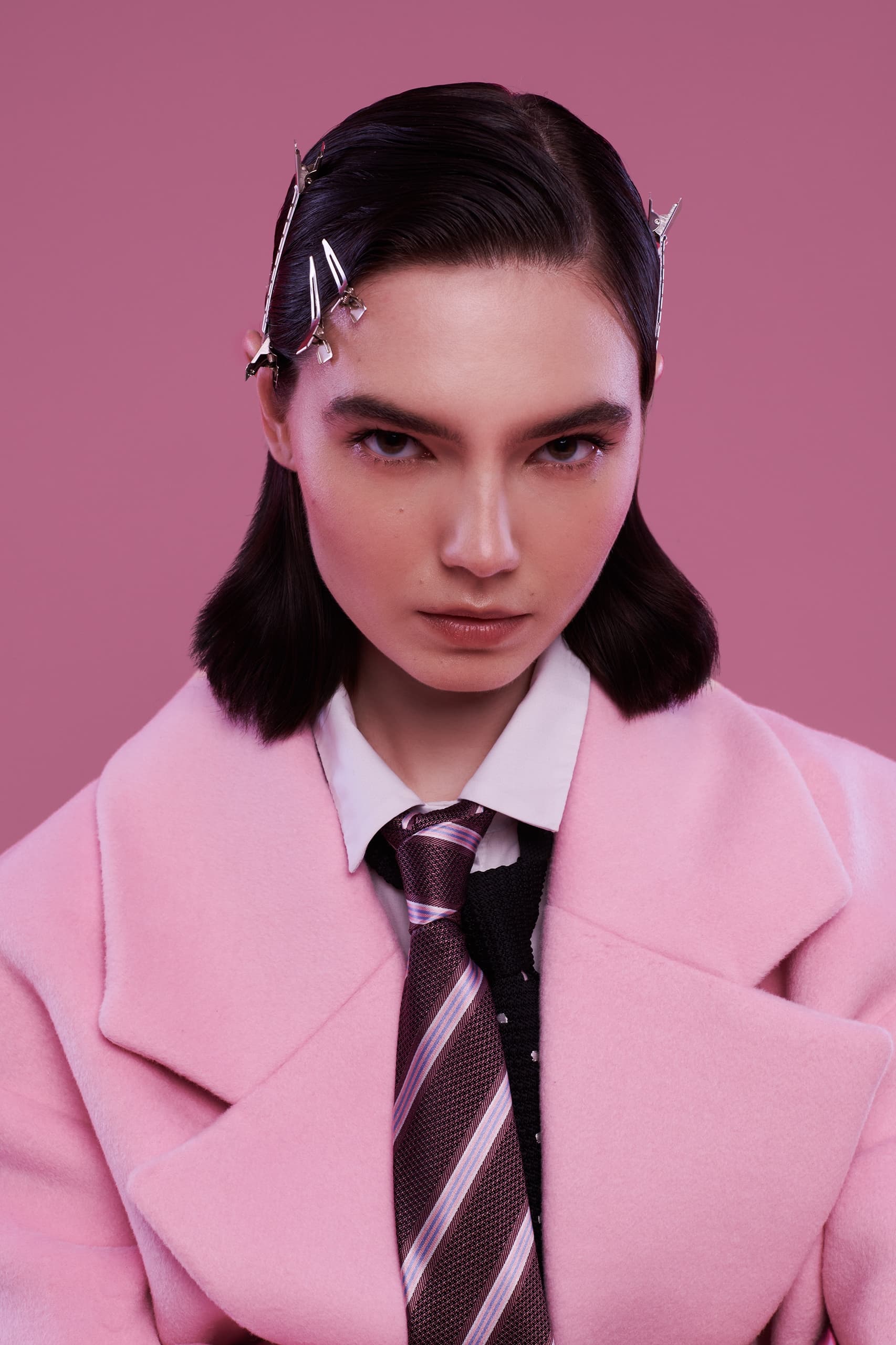 Fashionable person in a pink coat and tie, hair styled with clips on pink background.