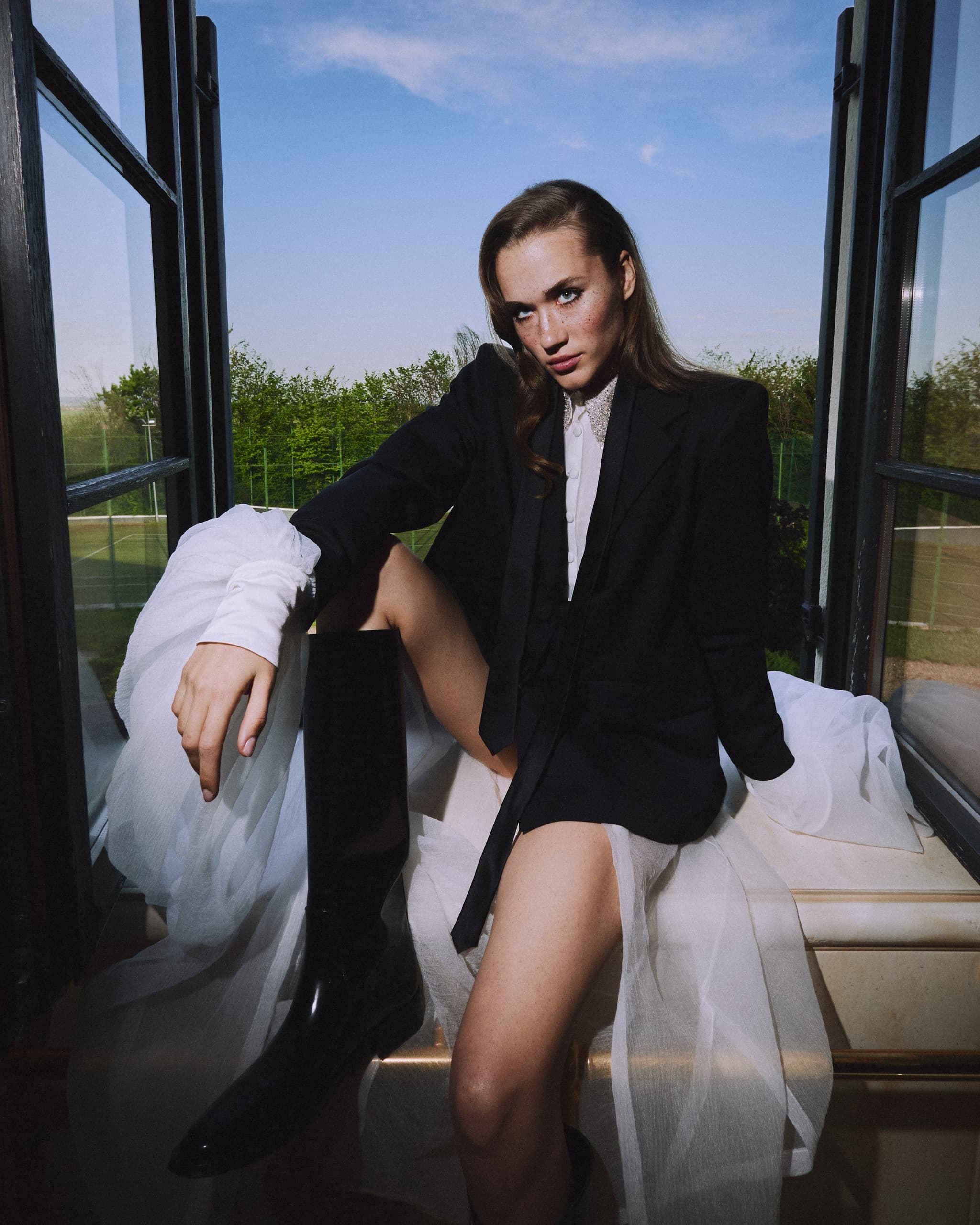 Fashionable woman in suit posing by open window with scenic outdoor view.