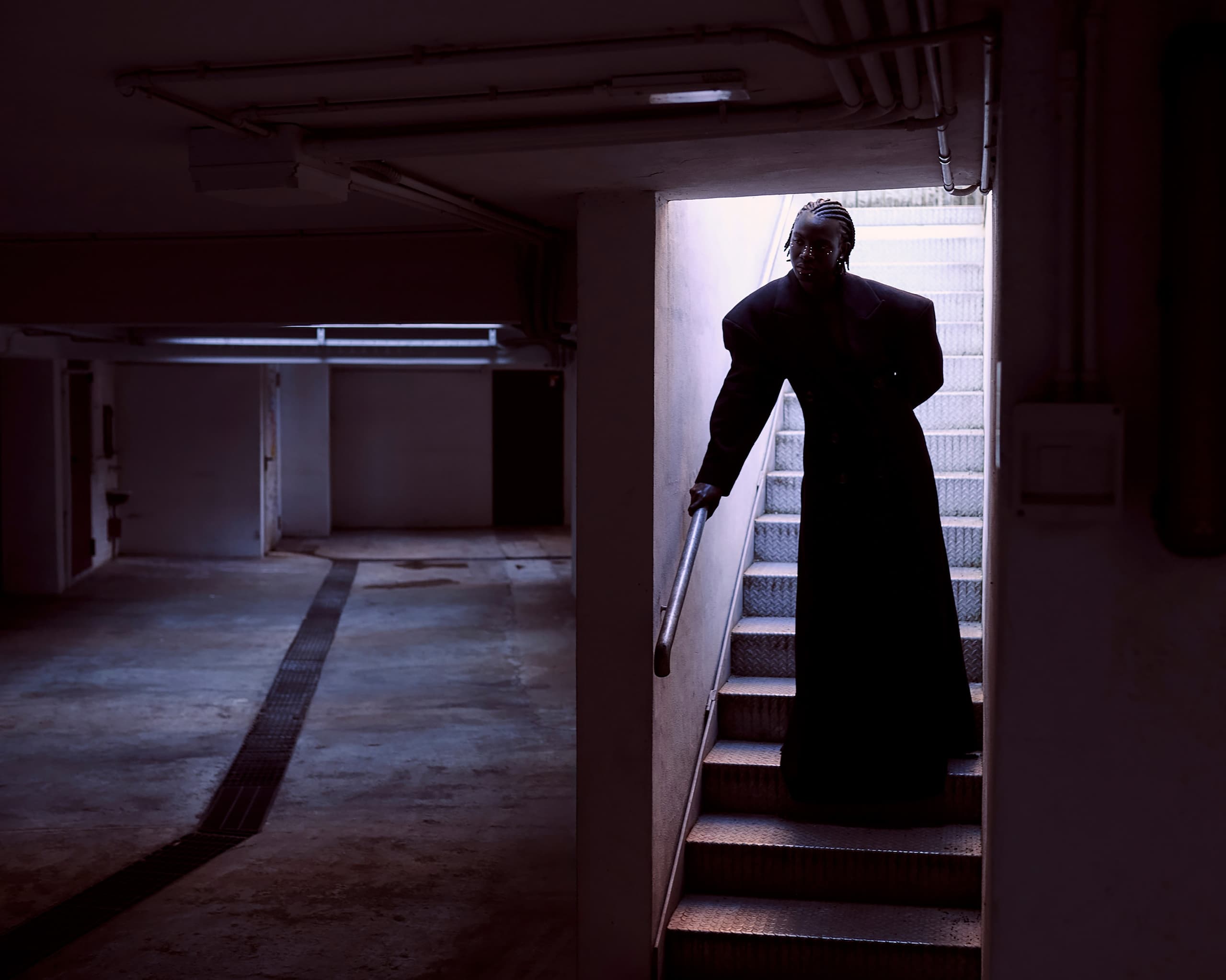 Person silhouetted in dark stairway, wearing long coat in shadowy basement setting.