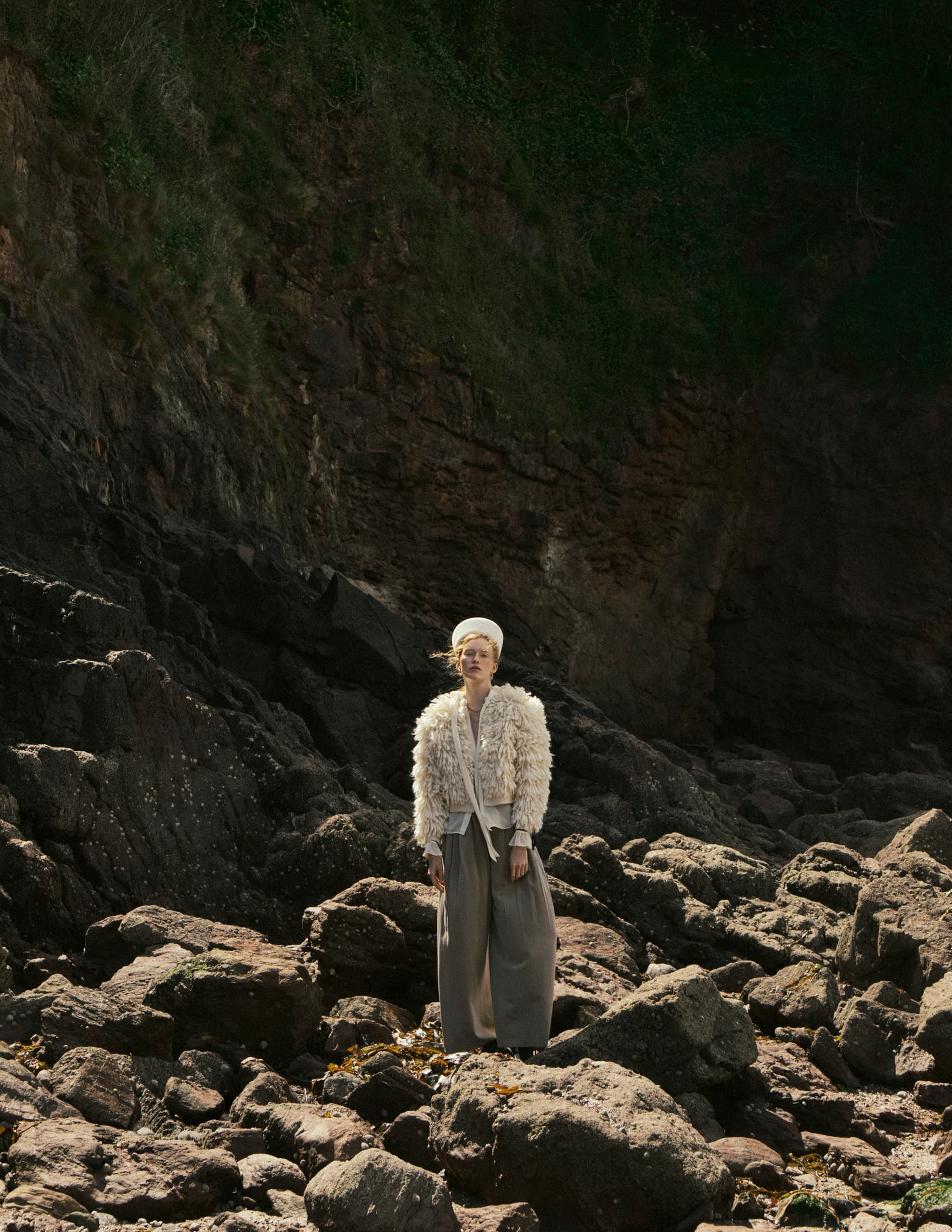 Person in stylish outfit stands on rocky coastline.