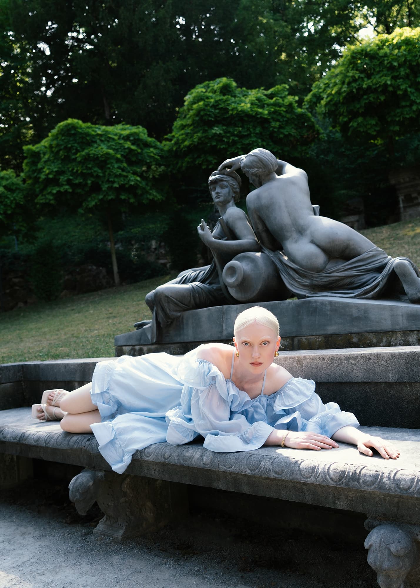 Woman in blue dress reclines on a stone bench beside classical sculptures in a park.