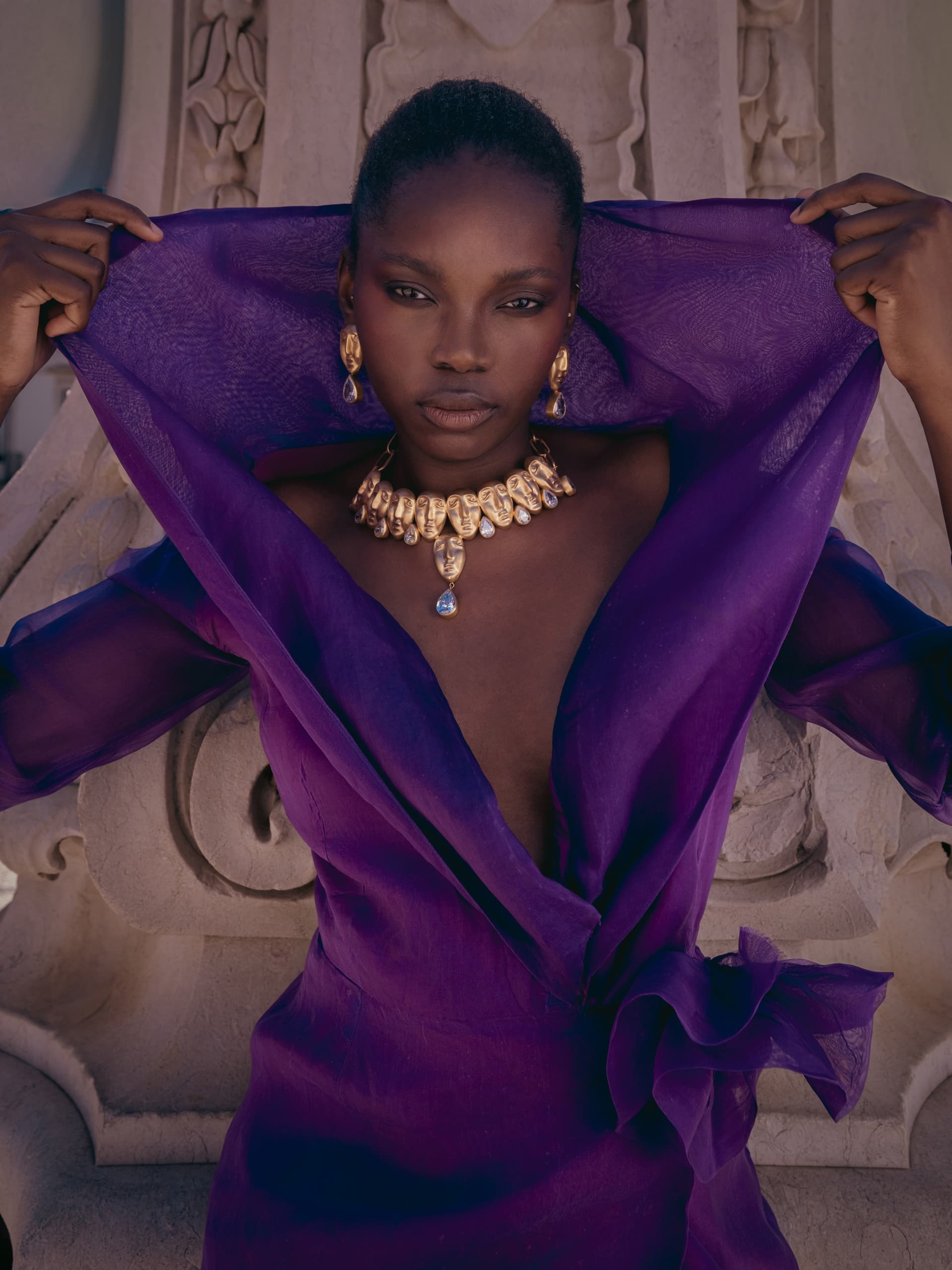 Person in elegant purple dress with gold jewelry, posing confidently outdoors.