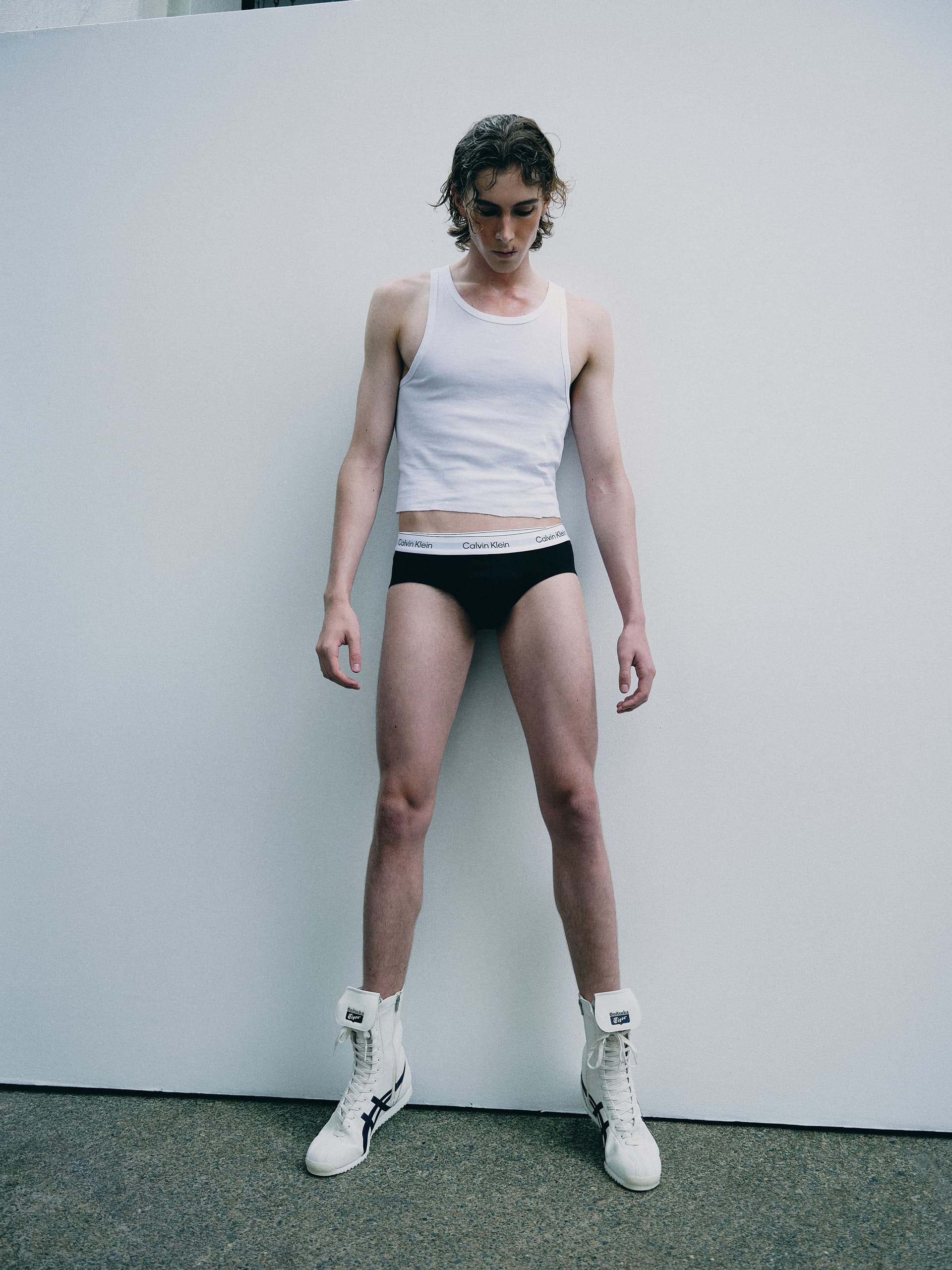 Model in white tank and black briefs posing against a minimalist backdrop.