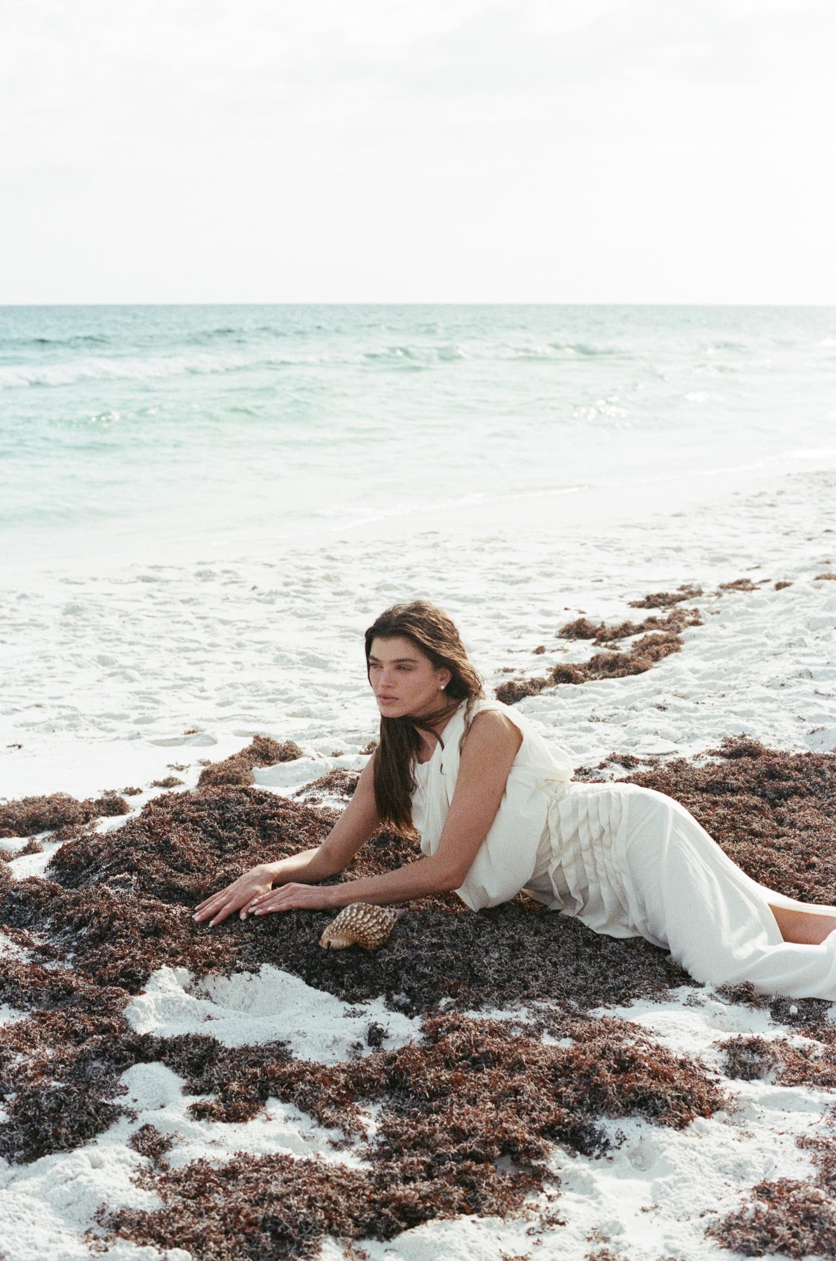 Woman in white dress lying on sandy beach with seaweed, ocean waves in background.