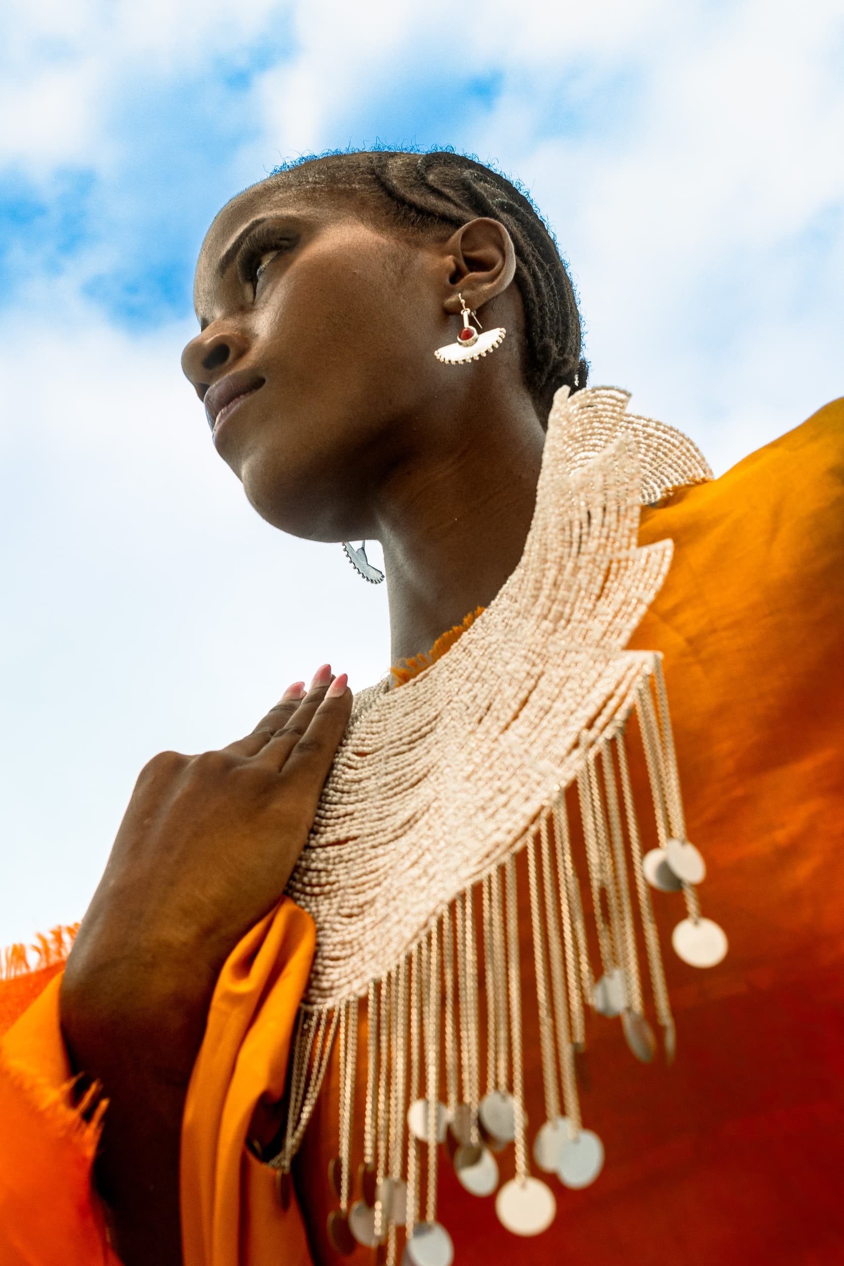 Woman in vibrant outfit with beaded necklace, looking upwards against blue sky.