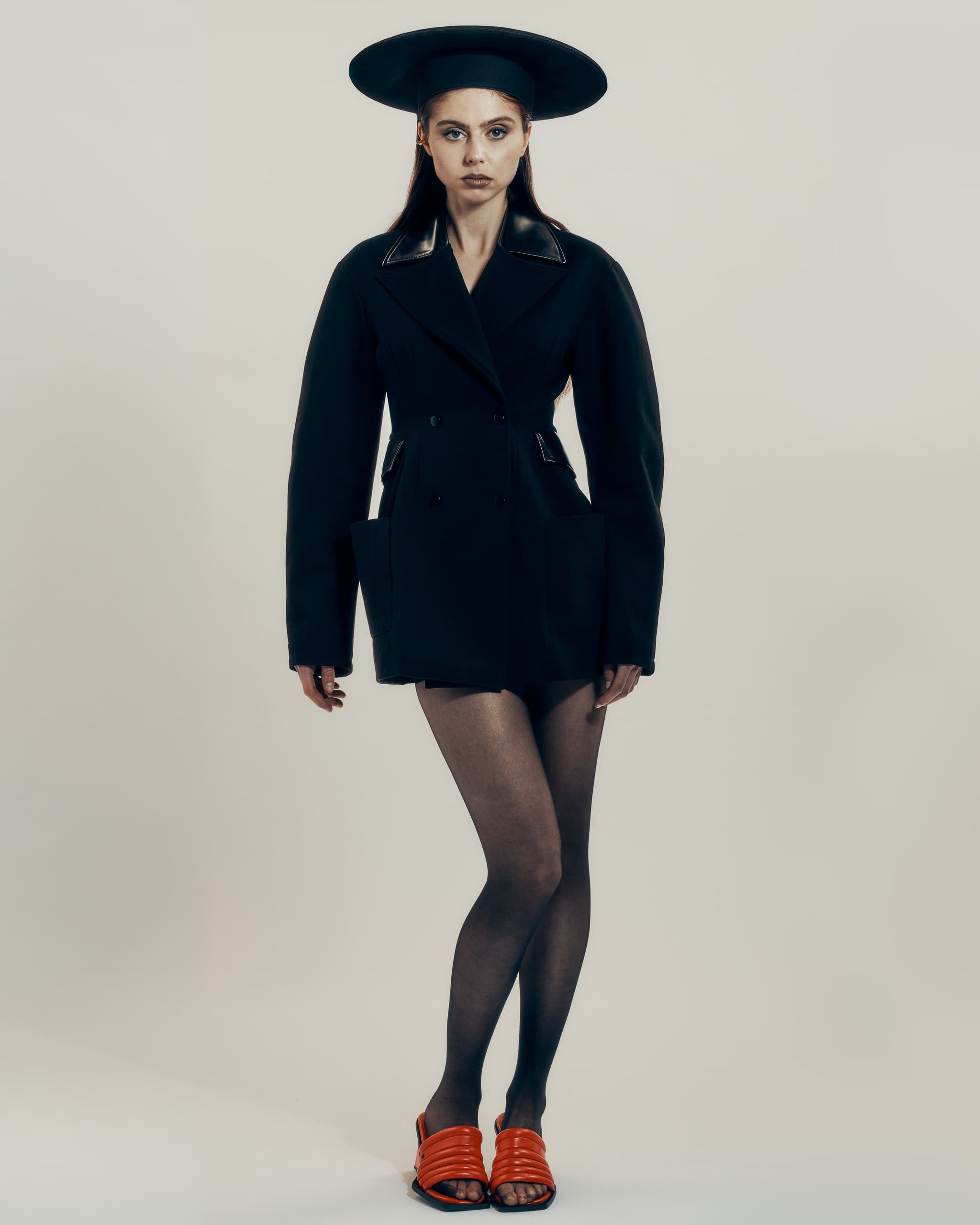 Fashion model in a black outfit, wide-brimmed hat, and red slides on a neutral background.