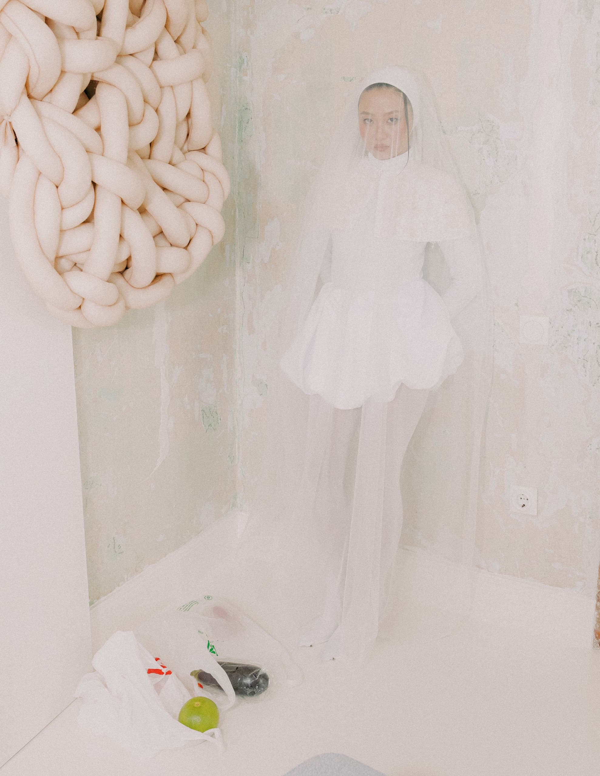 Person in avant-garde white dress with veil, next to abstract wall art and grocery bag.