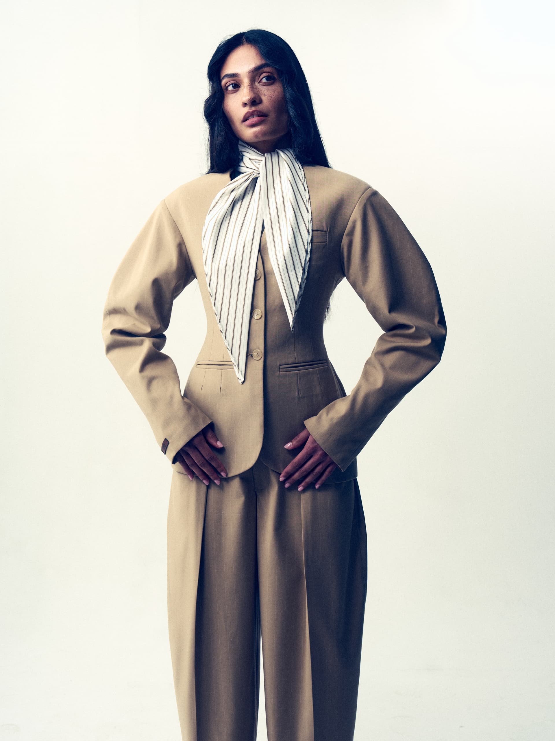 Woman in stylish beige suit with striped scarf, hands on hips, white background.