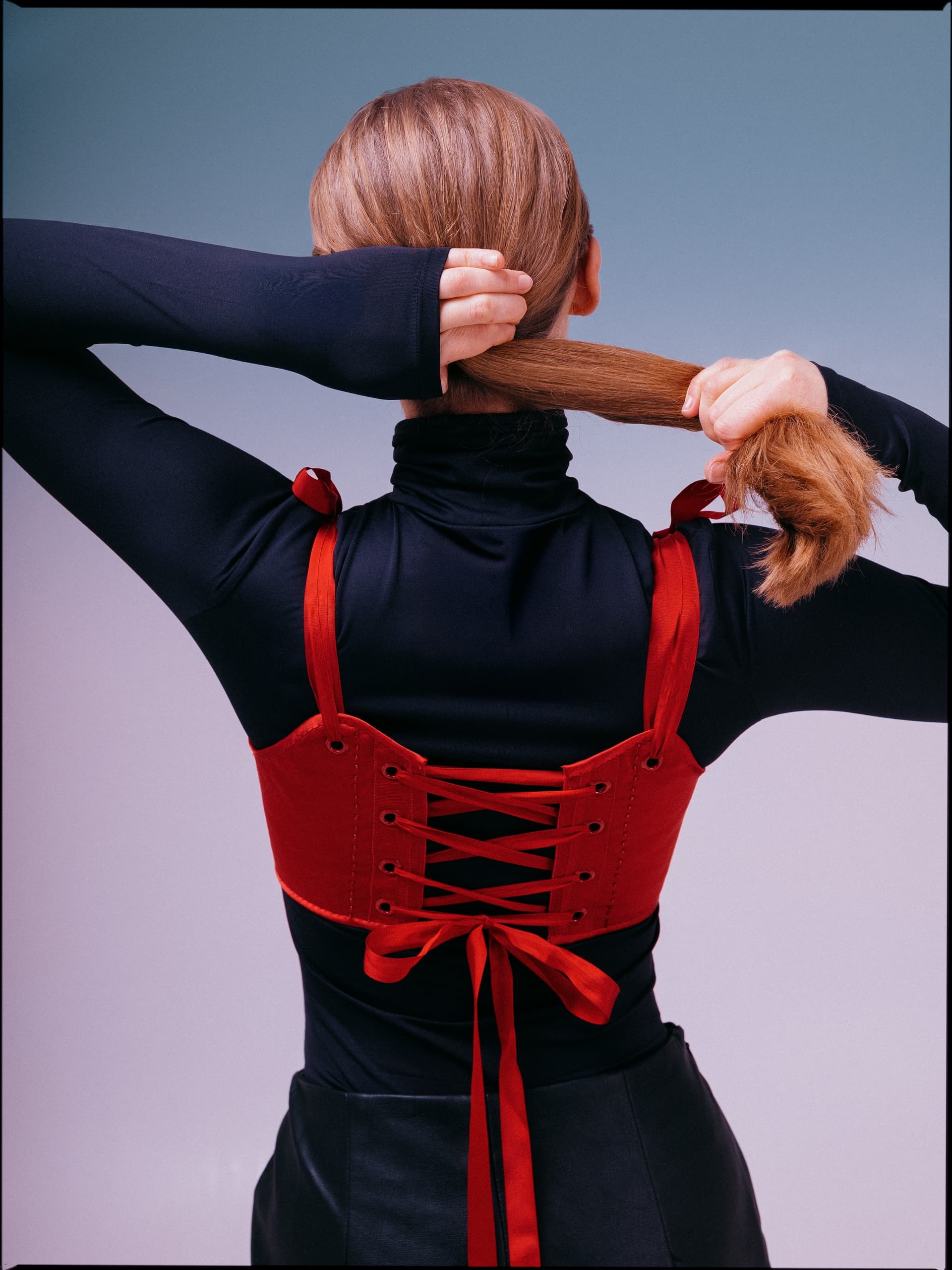 Woman in red lace-up corset tying hair, wearing black long-sleeve top.