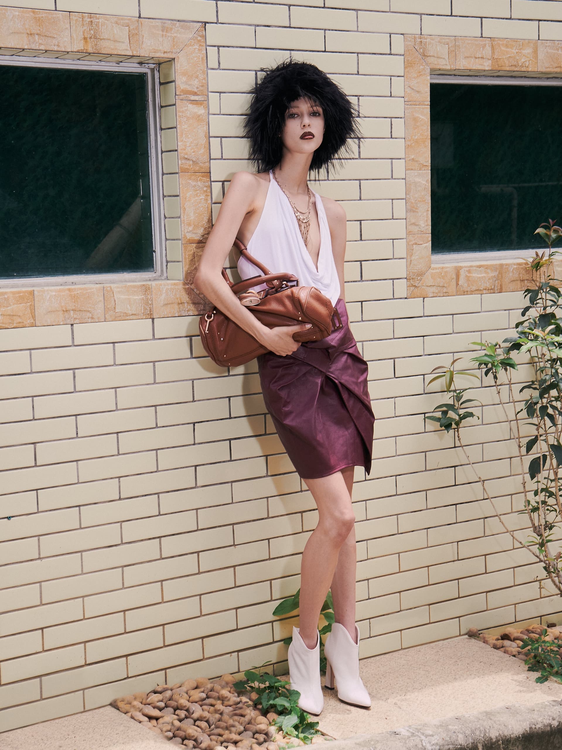 Woman in chic outfit holding leather bag, standing by a textured brick wall.
