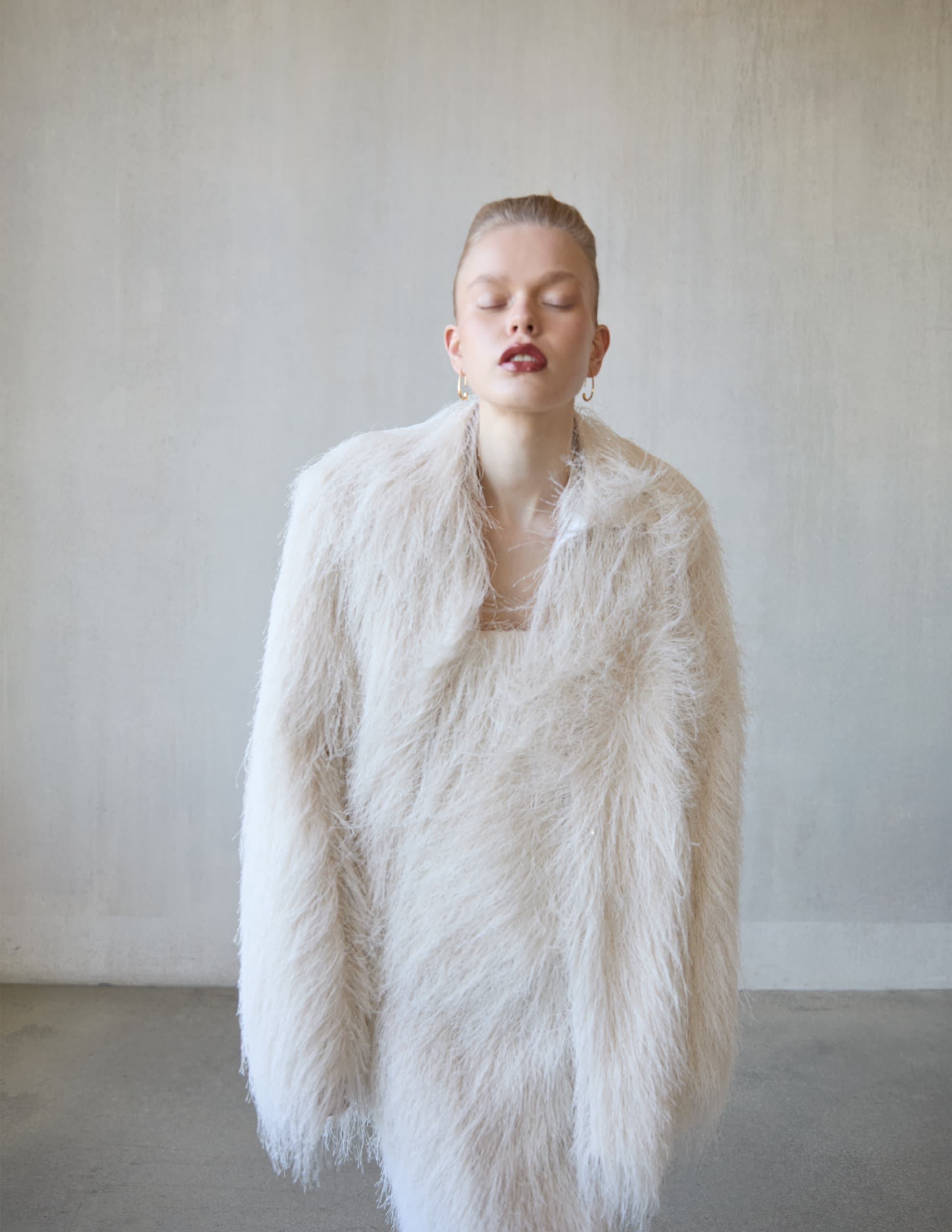Woman in fluffy white coat with eyes closed, minimalist background.