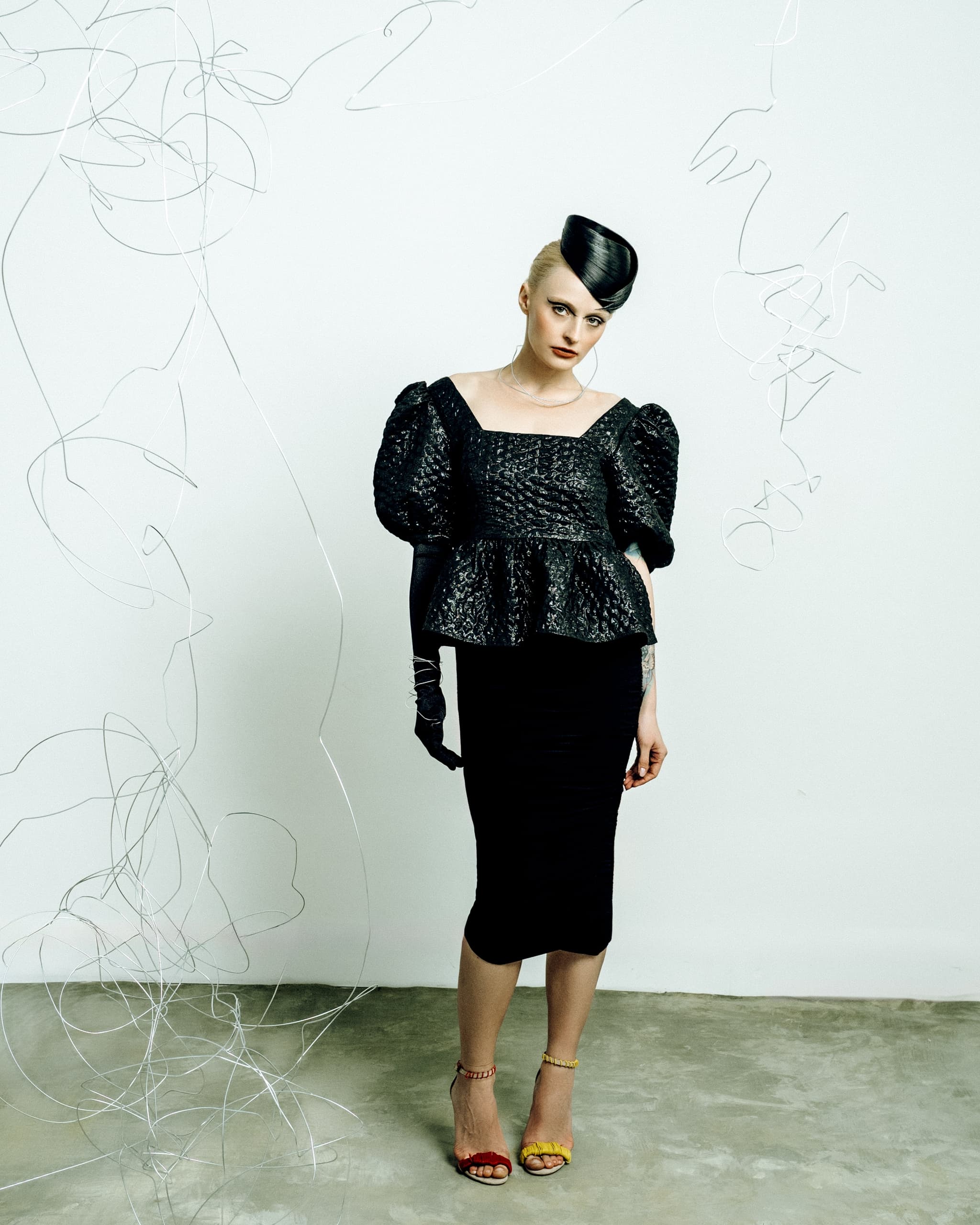 Modern fashion shoot with a model in black dress and hat, surrounded by wire art.