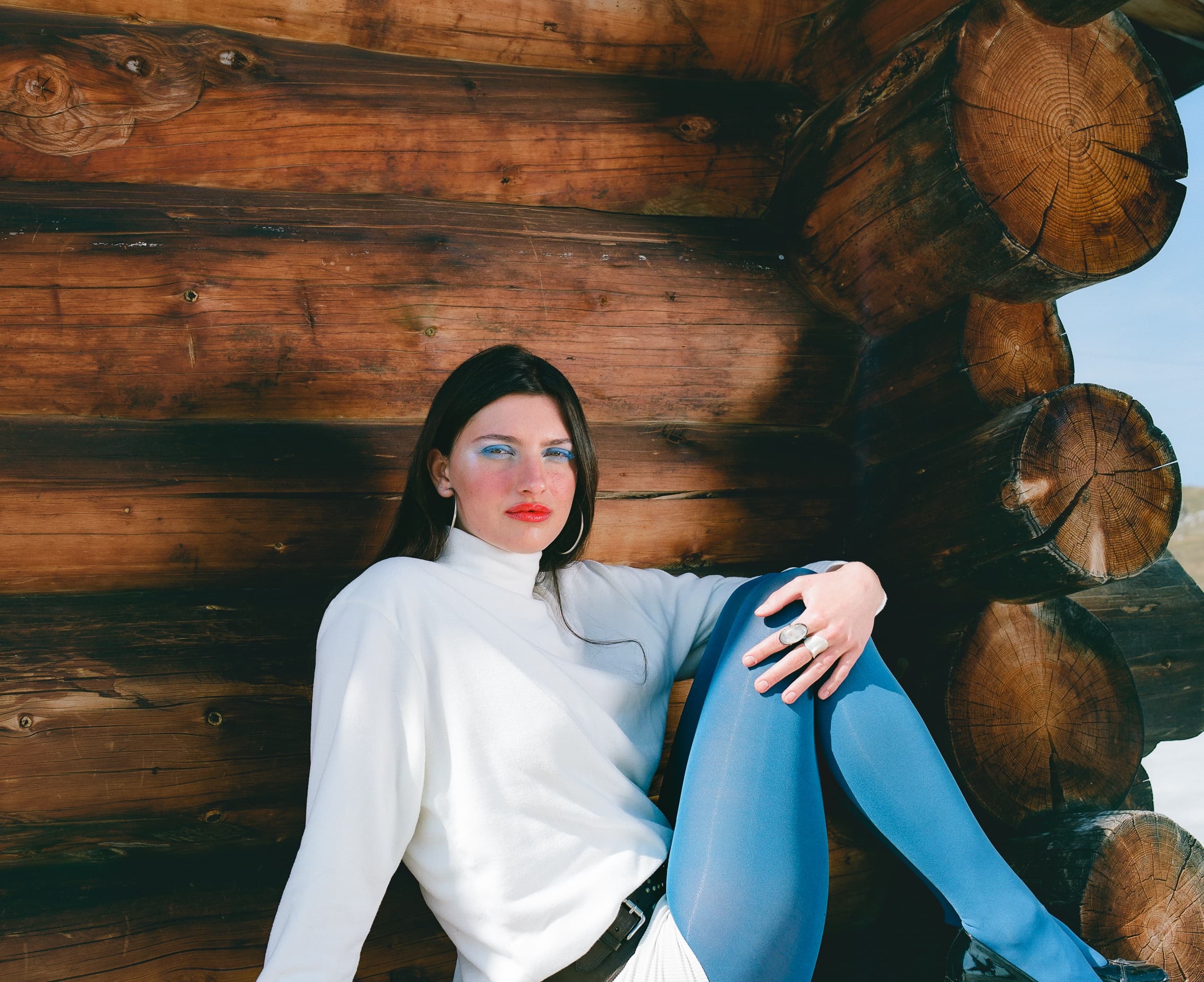 Woman in winter fashion, leaning on wood logs, with a rustic background.