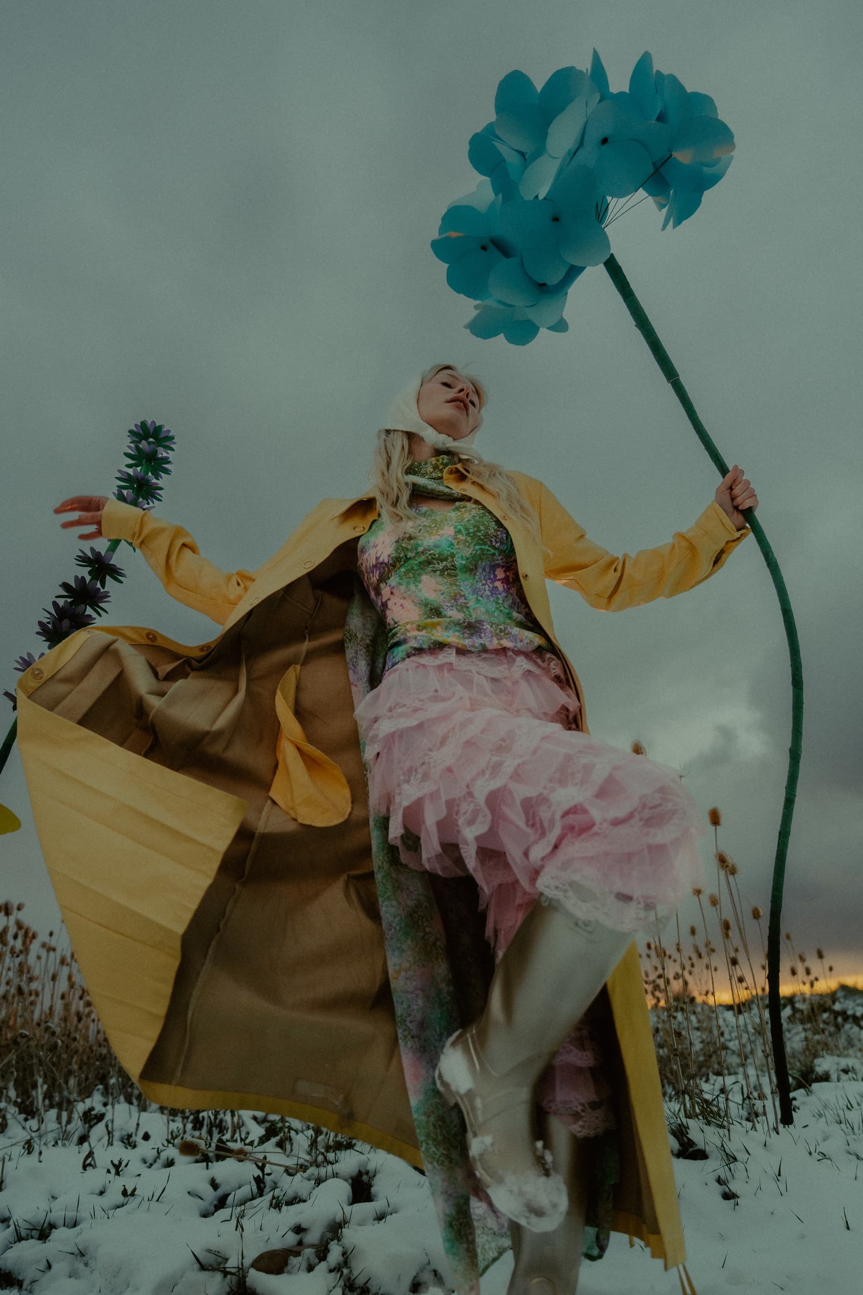 Stylish woman in colorful outfit holding large blue flowers in a snowy field.