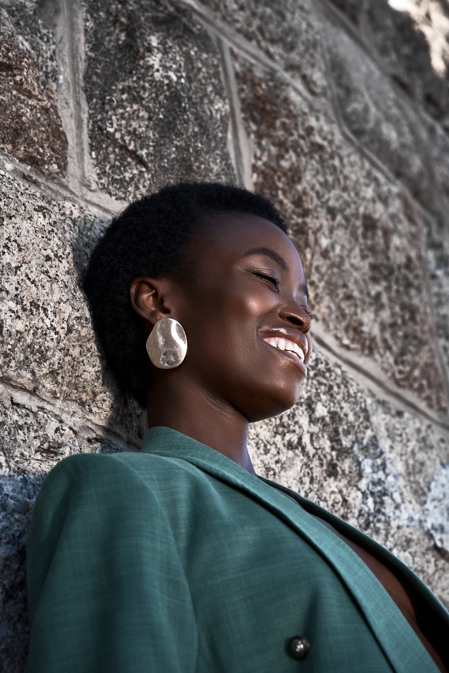 Smiling woman in green blazer leaning against stone wall, wearing large silver earrings.