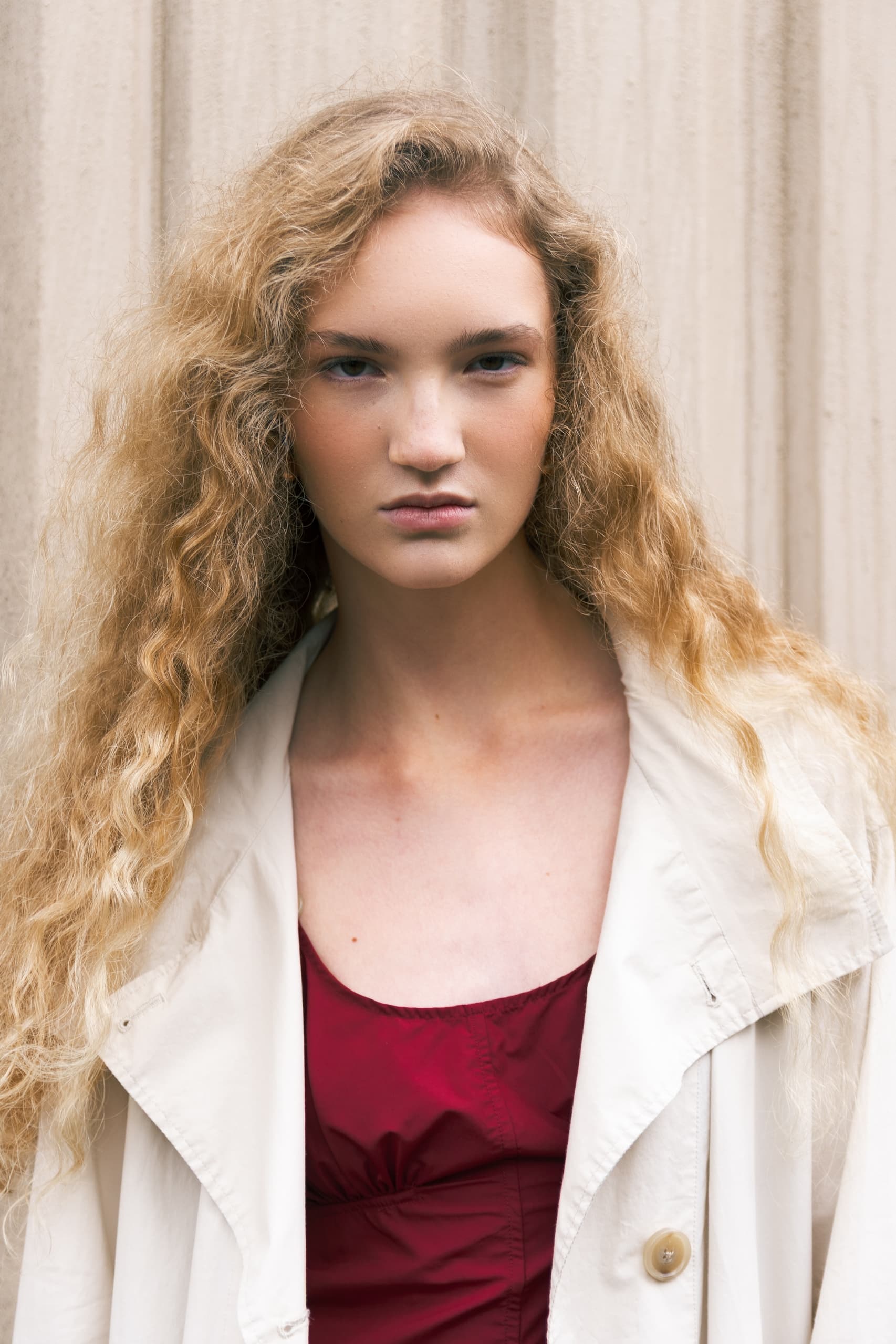 Young woman with curly hair in a red dress and beige jacket, neutral expression.