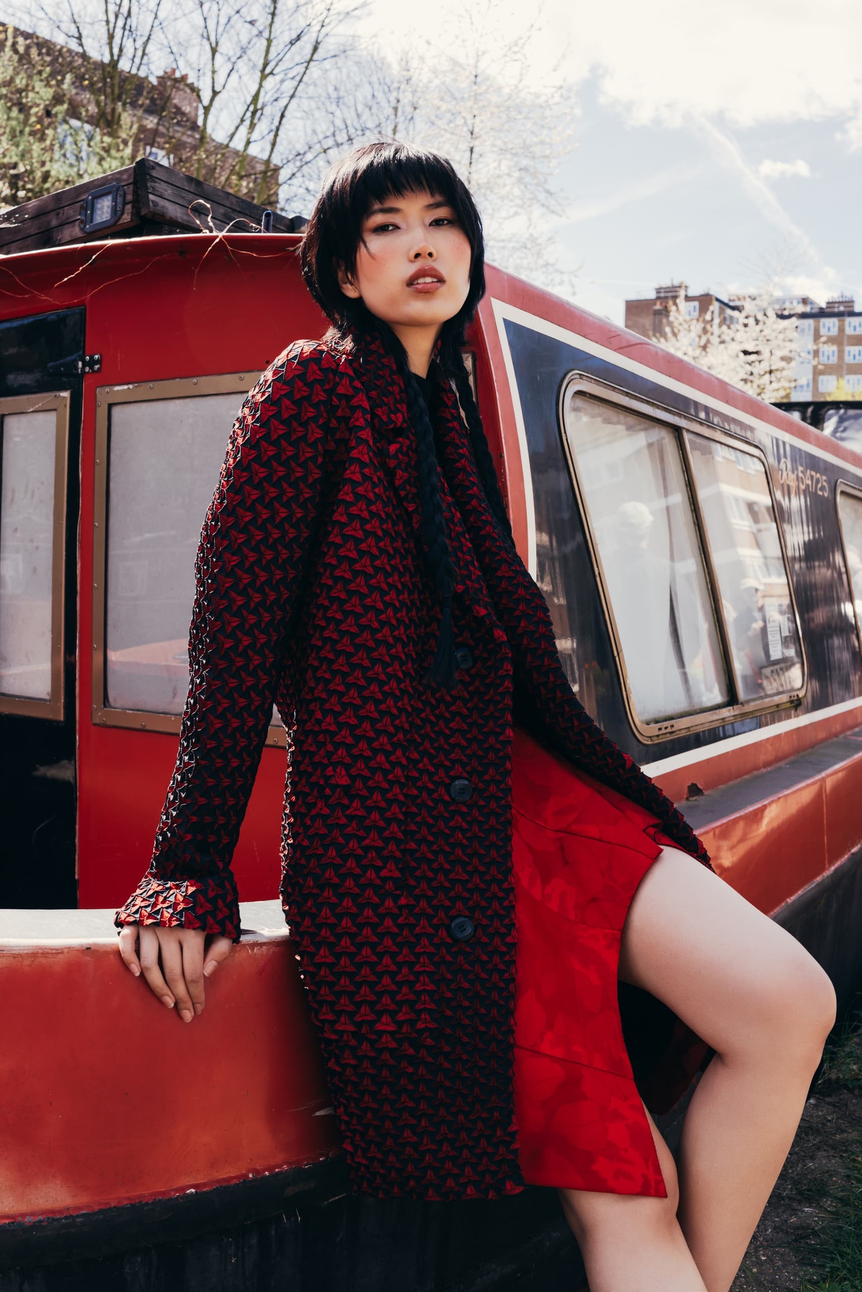 Fashionable woman in red coat leaning against red and black boat outdoors.
