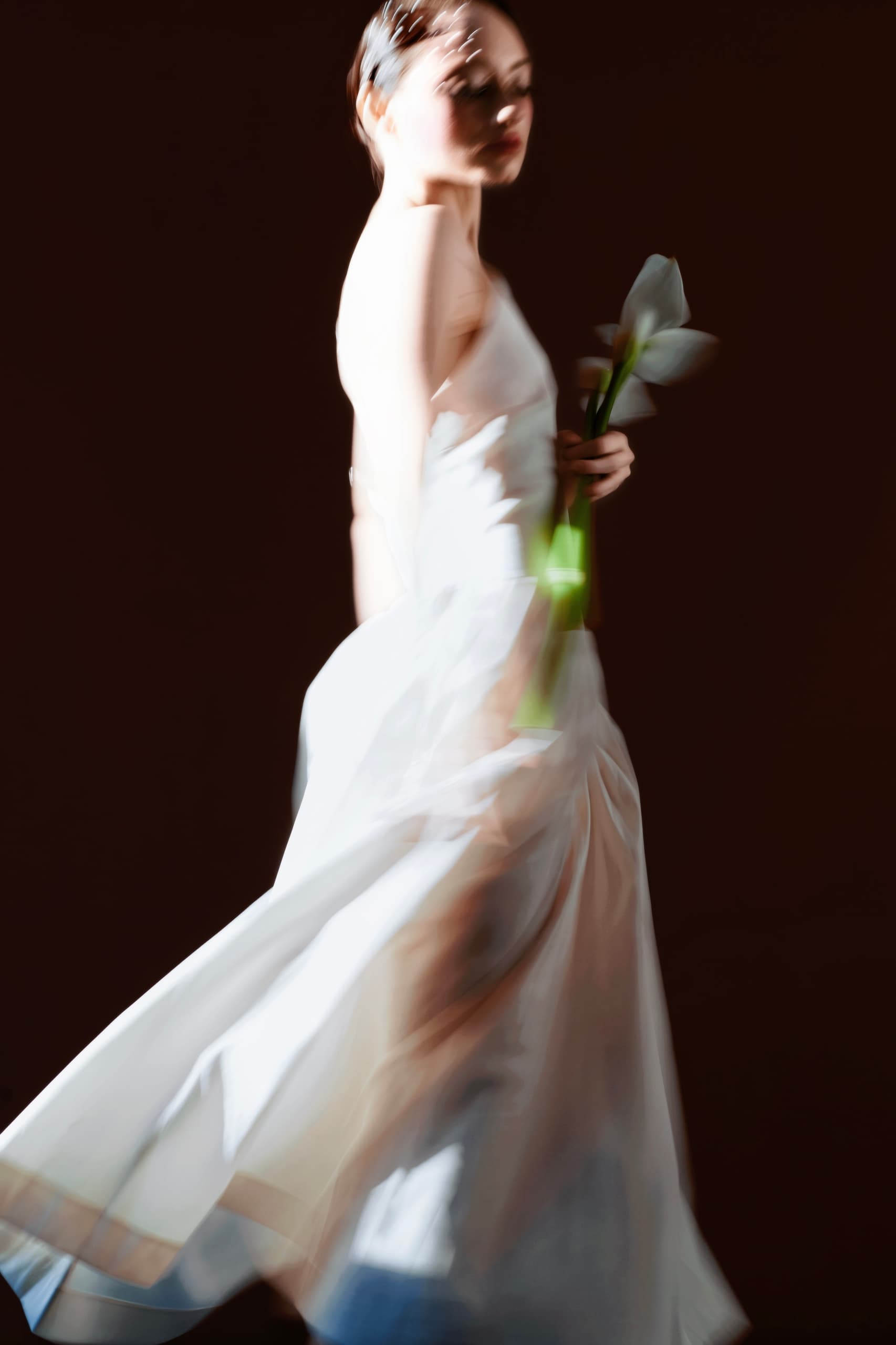 Blurred woman in flowing white dress holding a flower against a dark background.