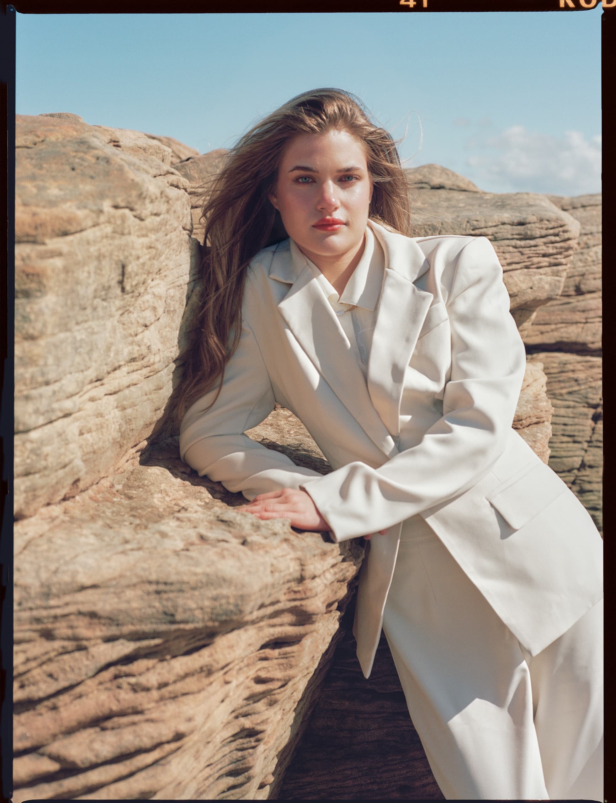 Woman in white suit leaning on scenic rock formation under sunny blue sky.