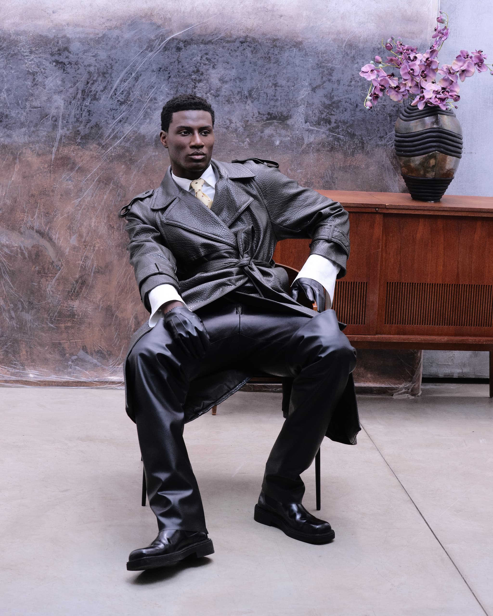 Stylish man in leather jacket sitting next to a wooden console and floral vase.