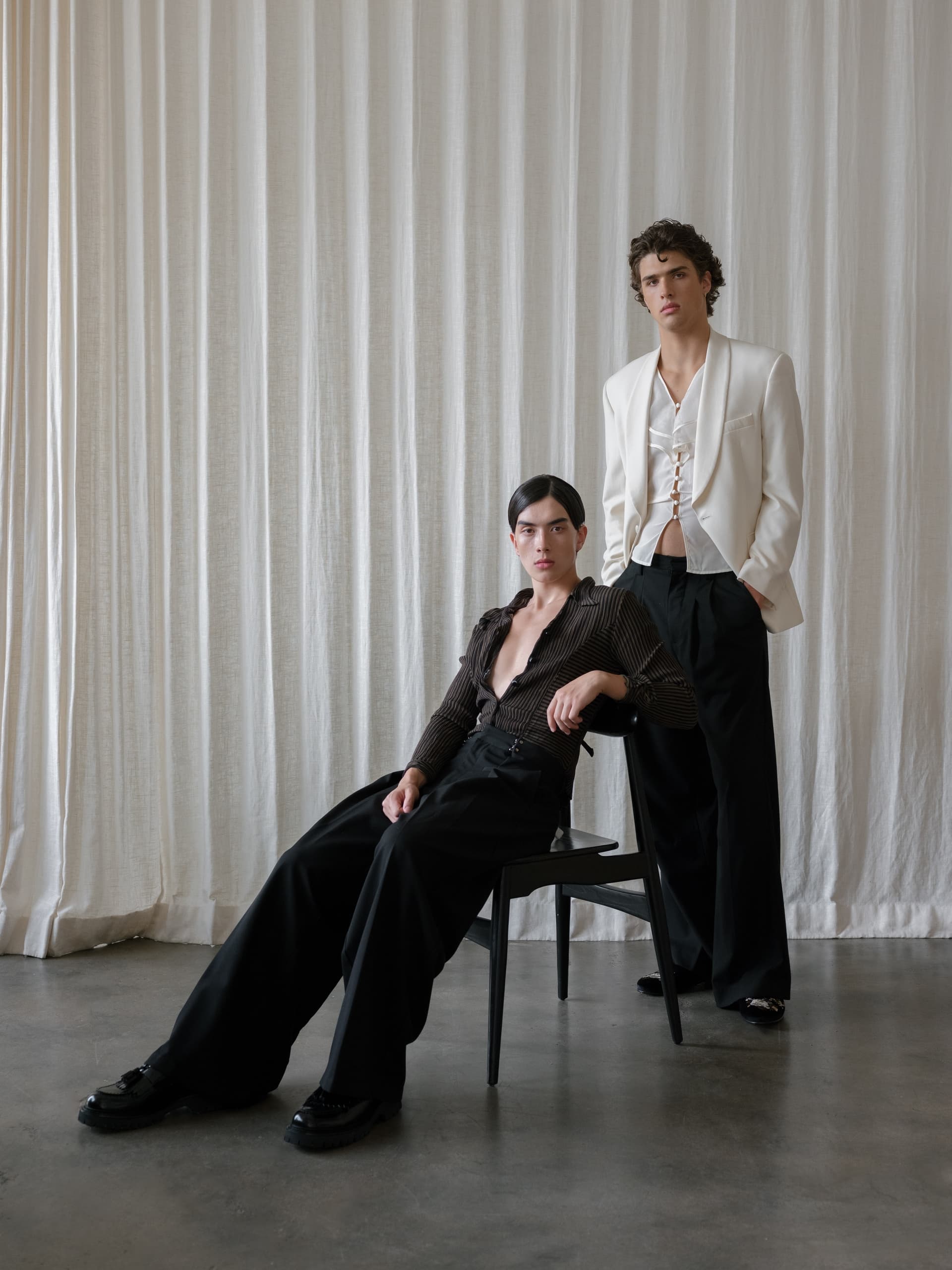 Two models in stylish outfits, sitting and standing against a white curtain backdrop.