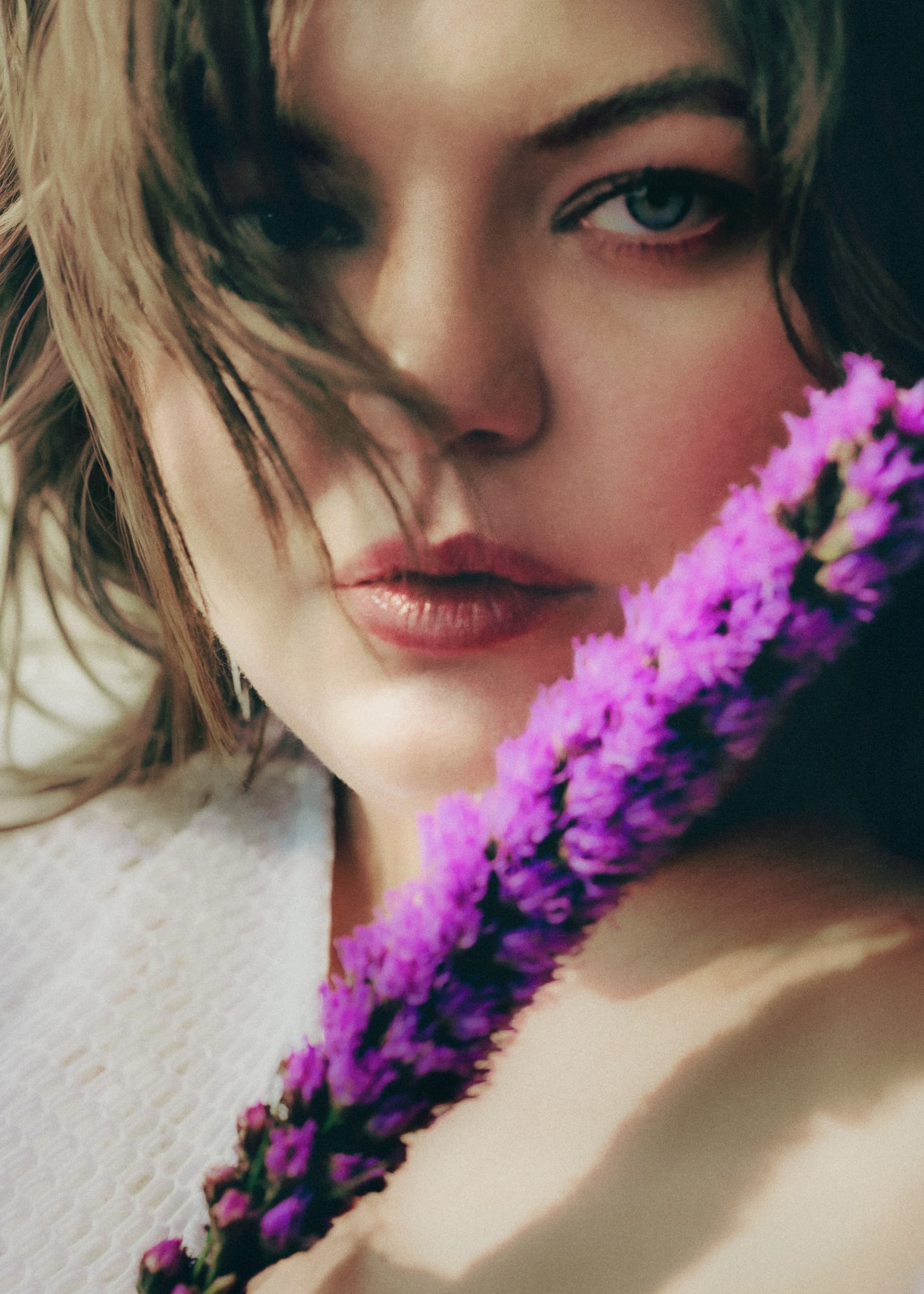 Close-up of a woman with purple flowers, focused expression, soft tones.