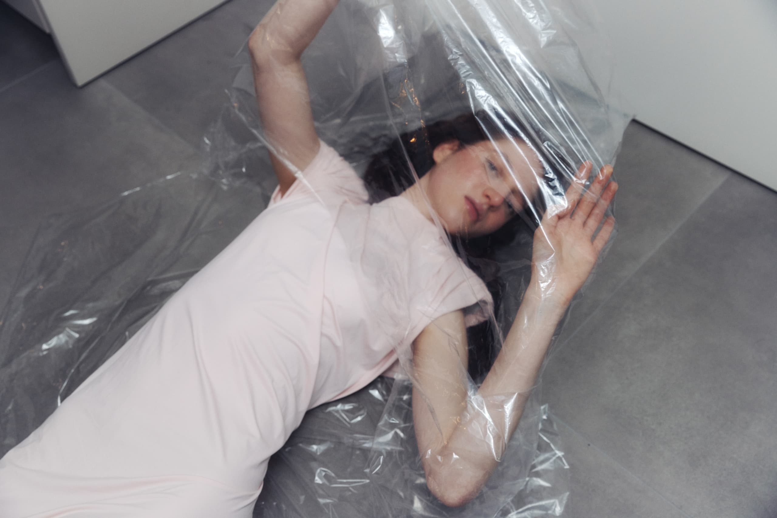 Person lying under clear plastic wrap on a floor in soft lighting.