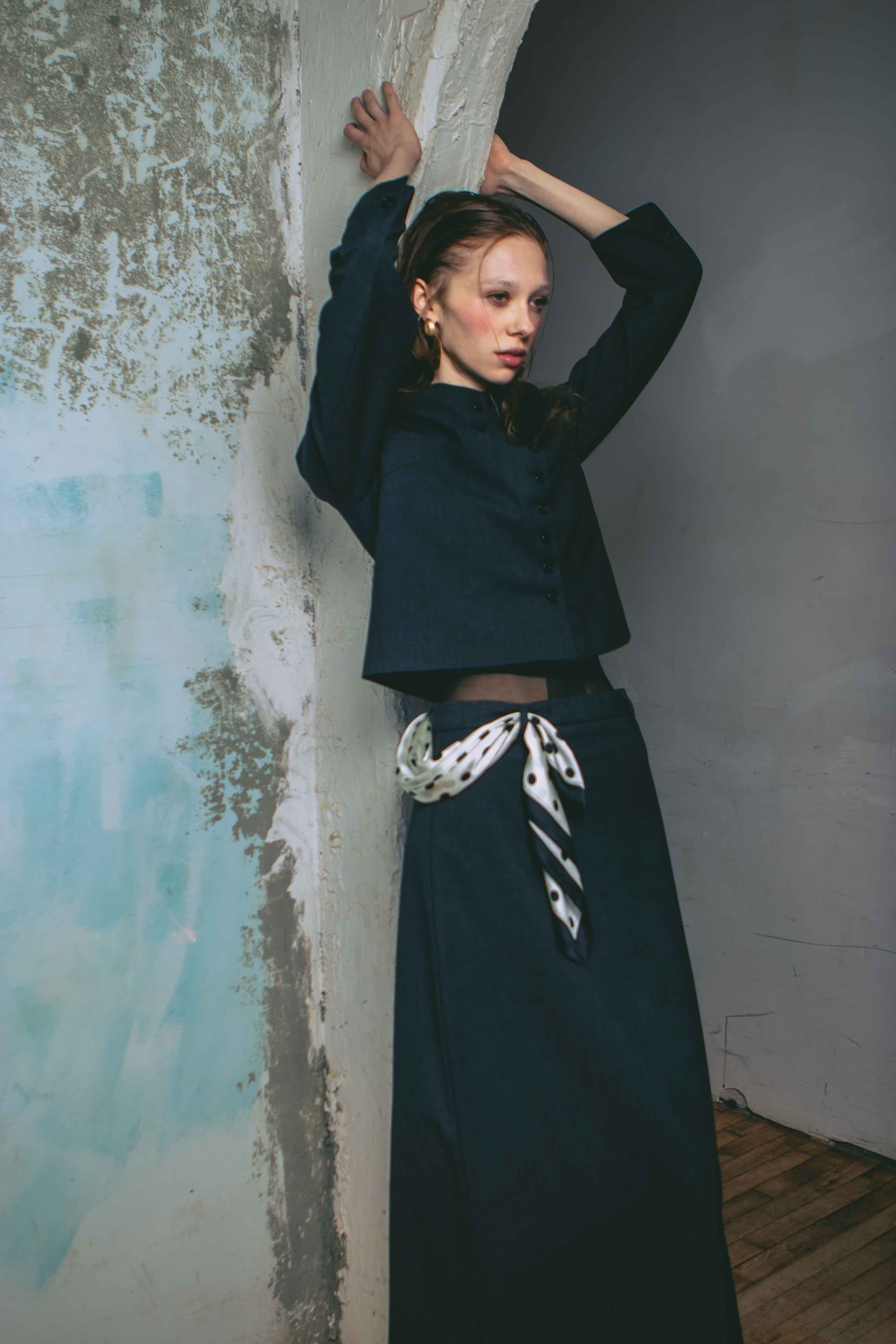 Fashionable woman in dark outfit leaning against distressed wall.