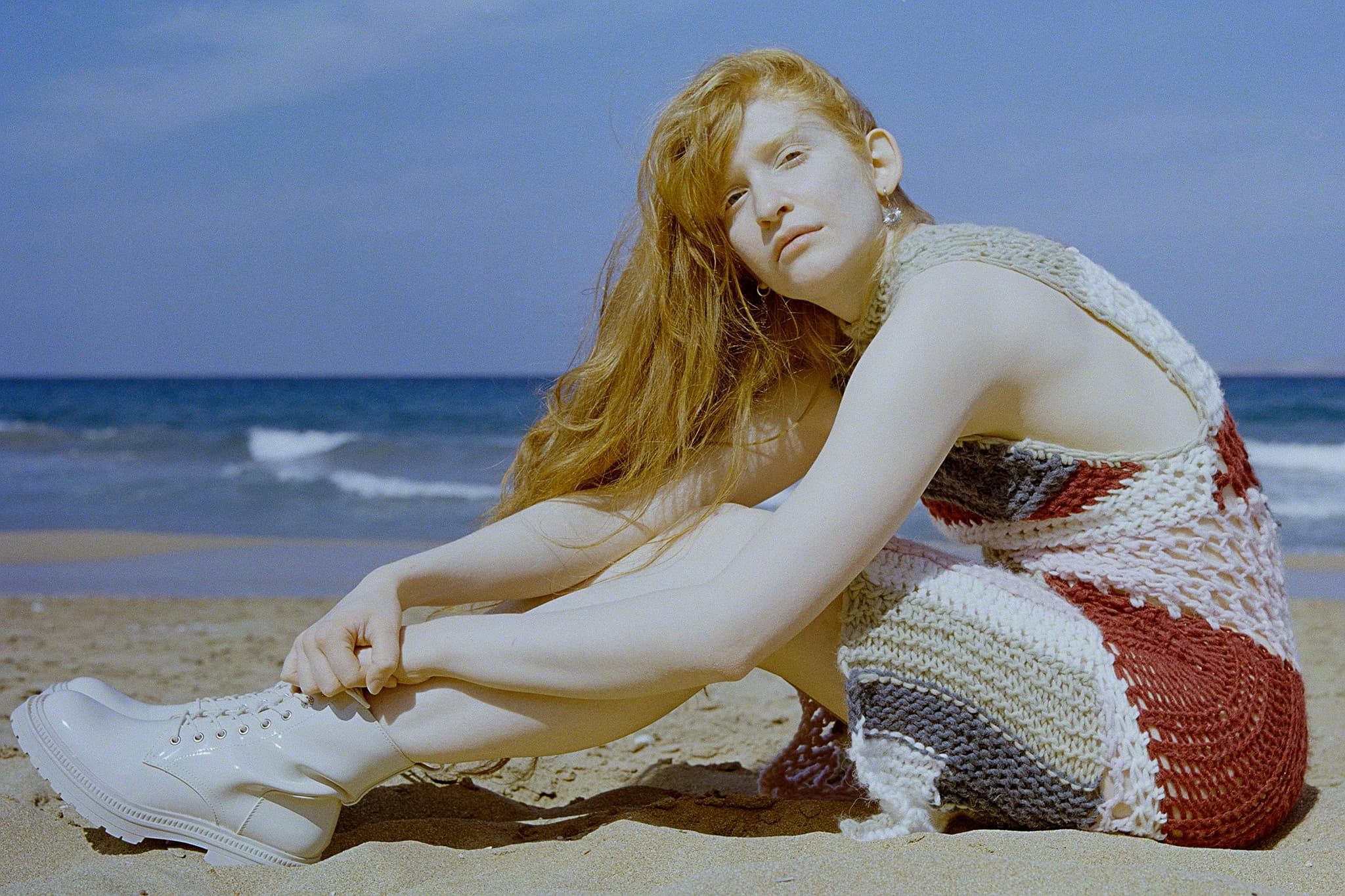 Woman in stylish knit dress sitting on the beach, white boots, serene sea background.