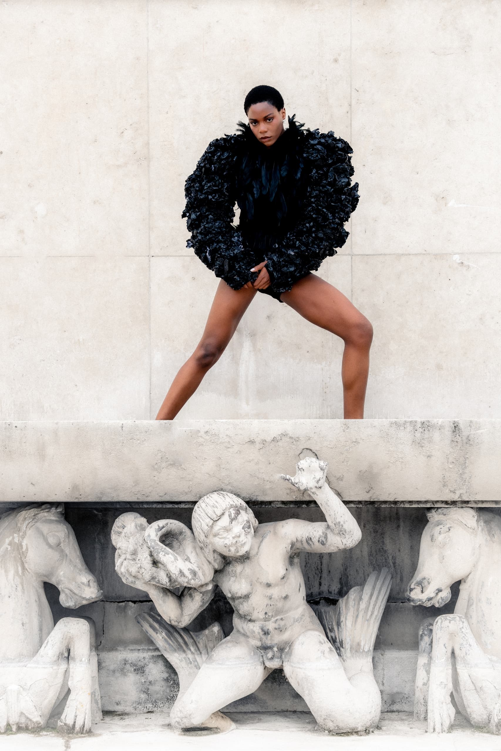 Person in black fashion attire posing above stone sculptures.