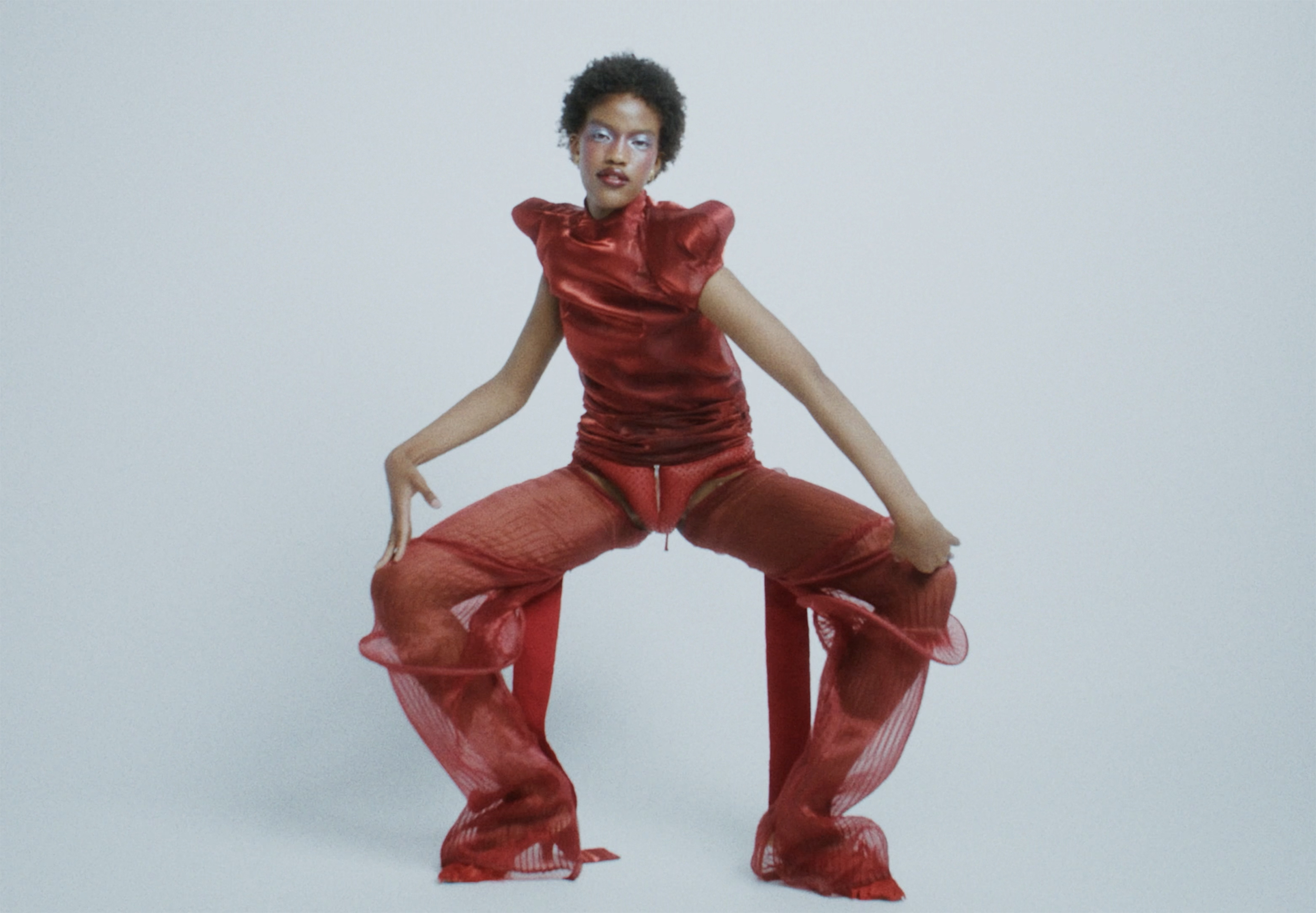 Fashion model in red sheer outfit posing in dynamic stance against plain backdrop.