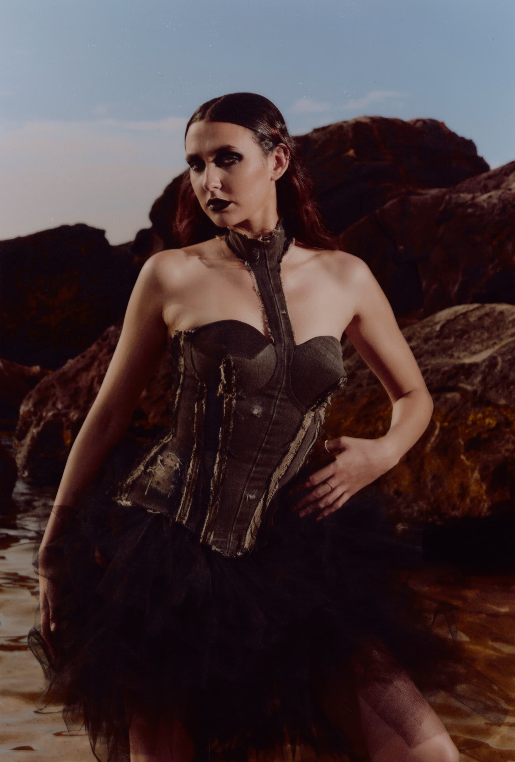 Gothic fashion model in a corset dress poses by rocks in moody lighting.