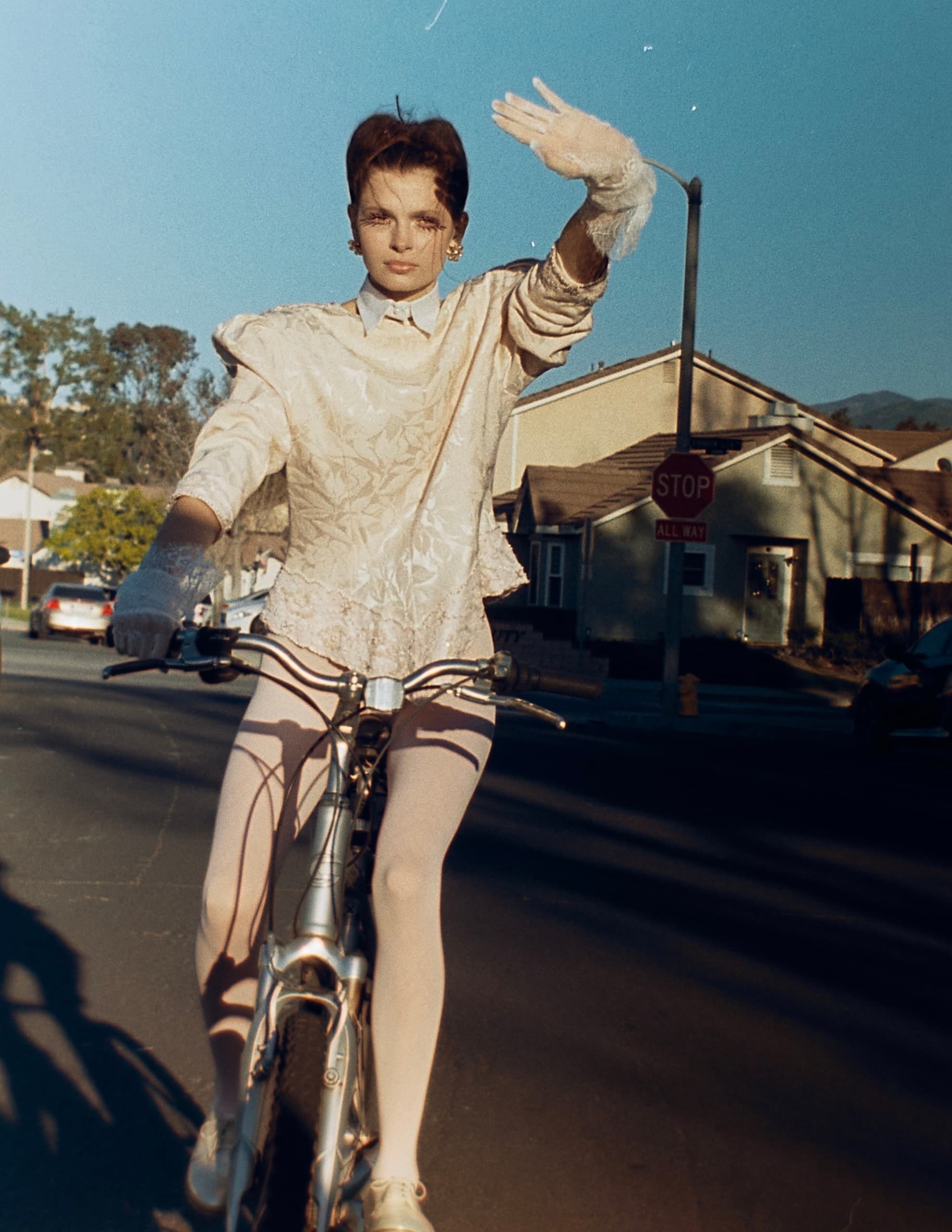 Person cycling in vintage attire on a suburban street, hand raised in sunlight.