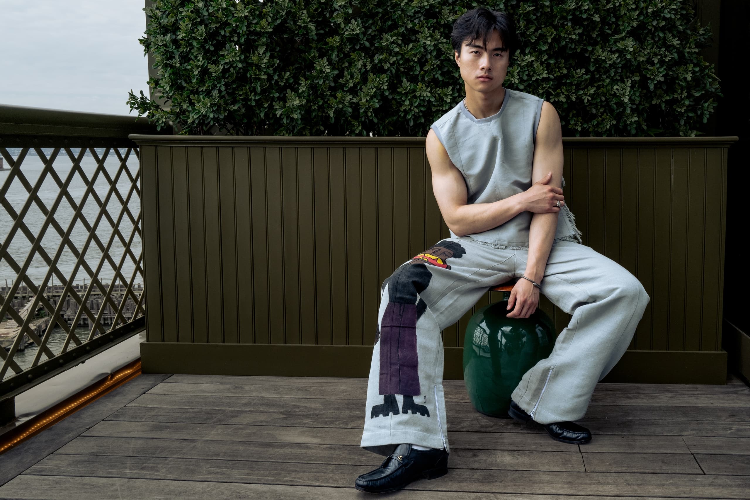 Man in stylish fashion posing on a deck with green plants and ocean view.