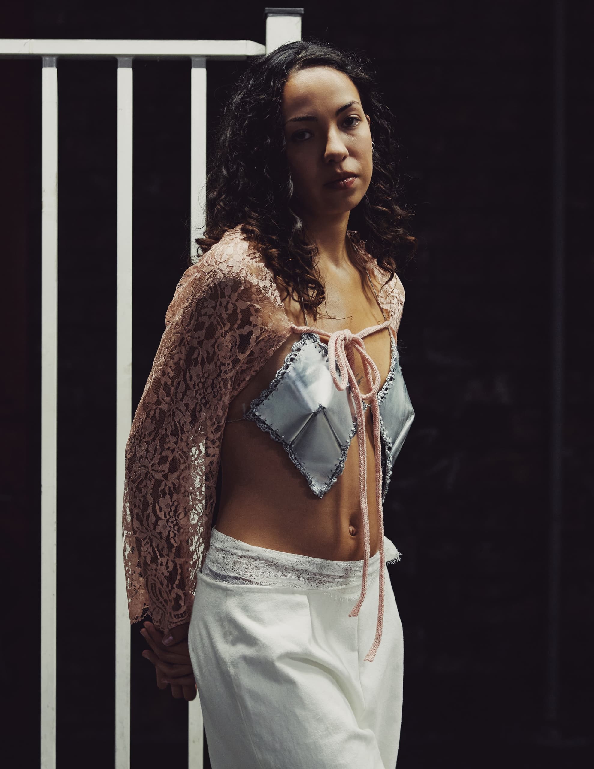 Woman in lace top and white pants standing by a white fence, in low light.