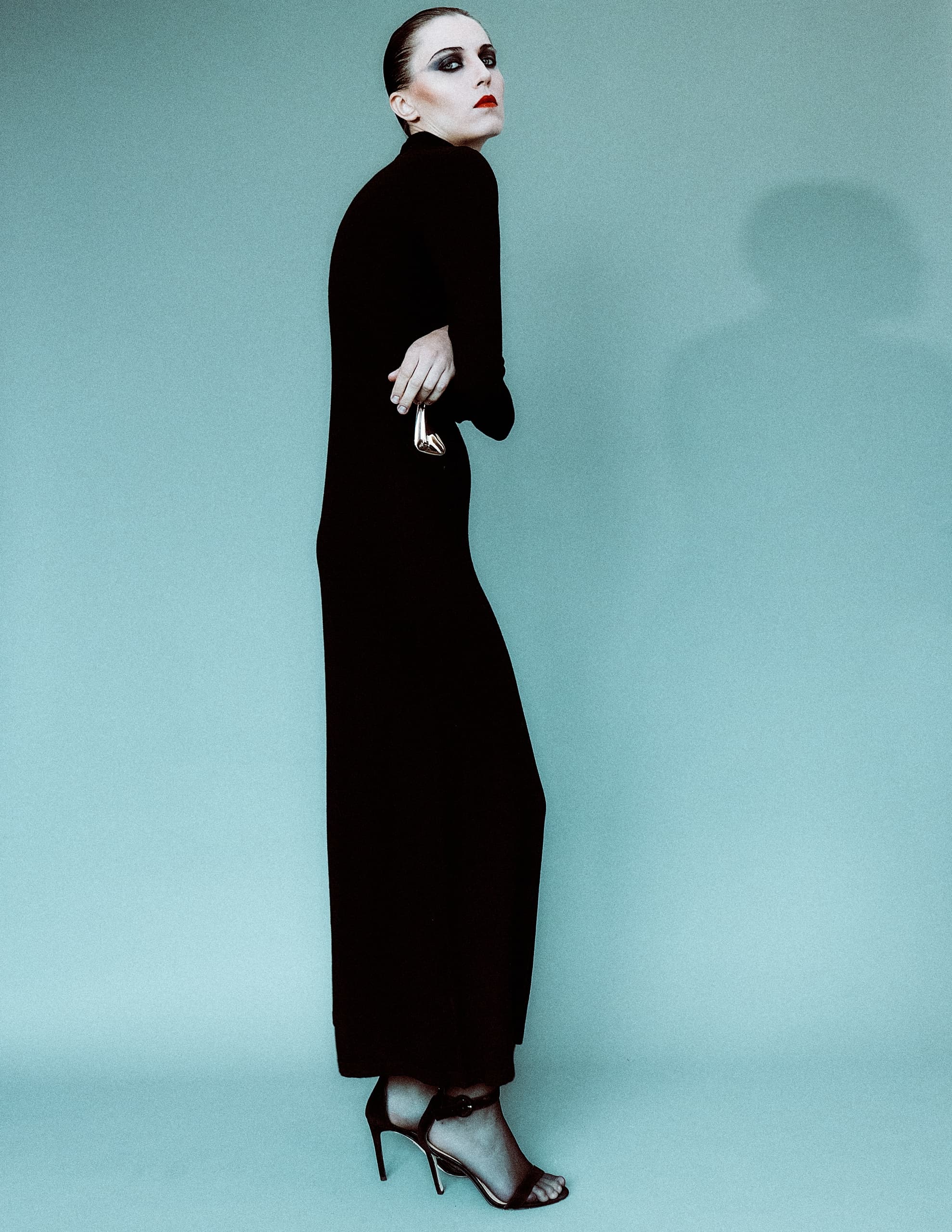 Fashion model in black dress and heels, striking pose against teal background.