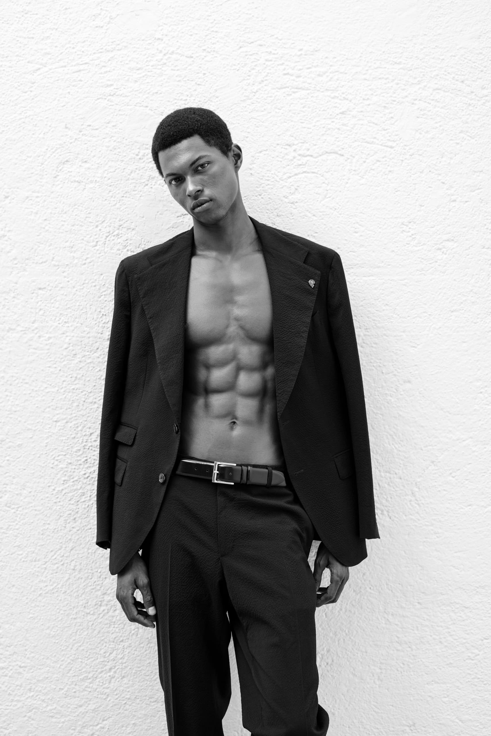 Man in black suit with open jacket, showing toned abs against a textured wall.