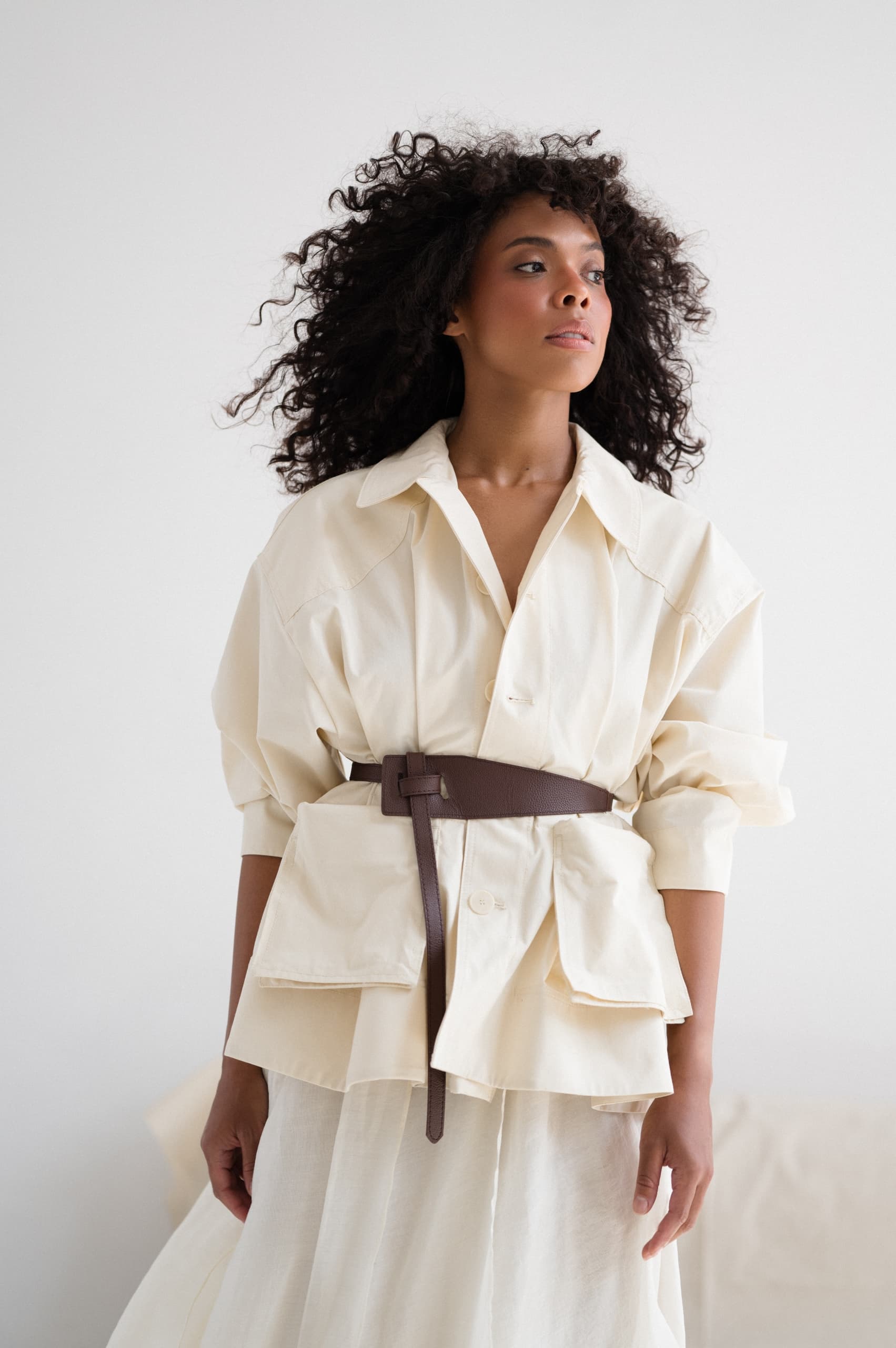 Woman in stylish cream outfit with belt, posing confidently against a minimalist backdrop.