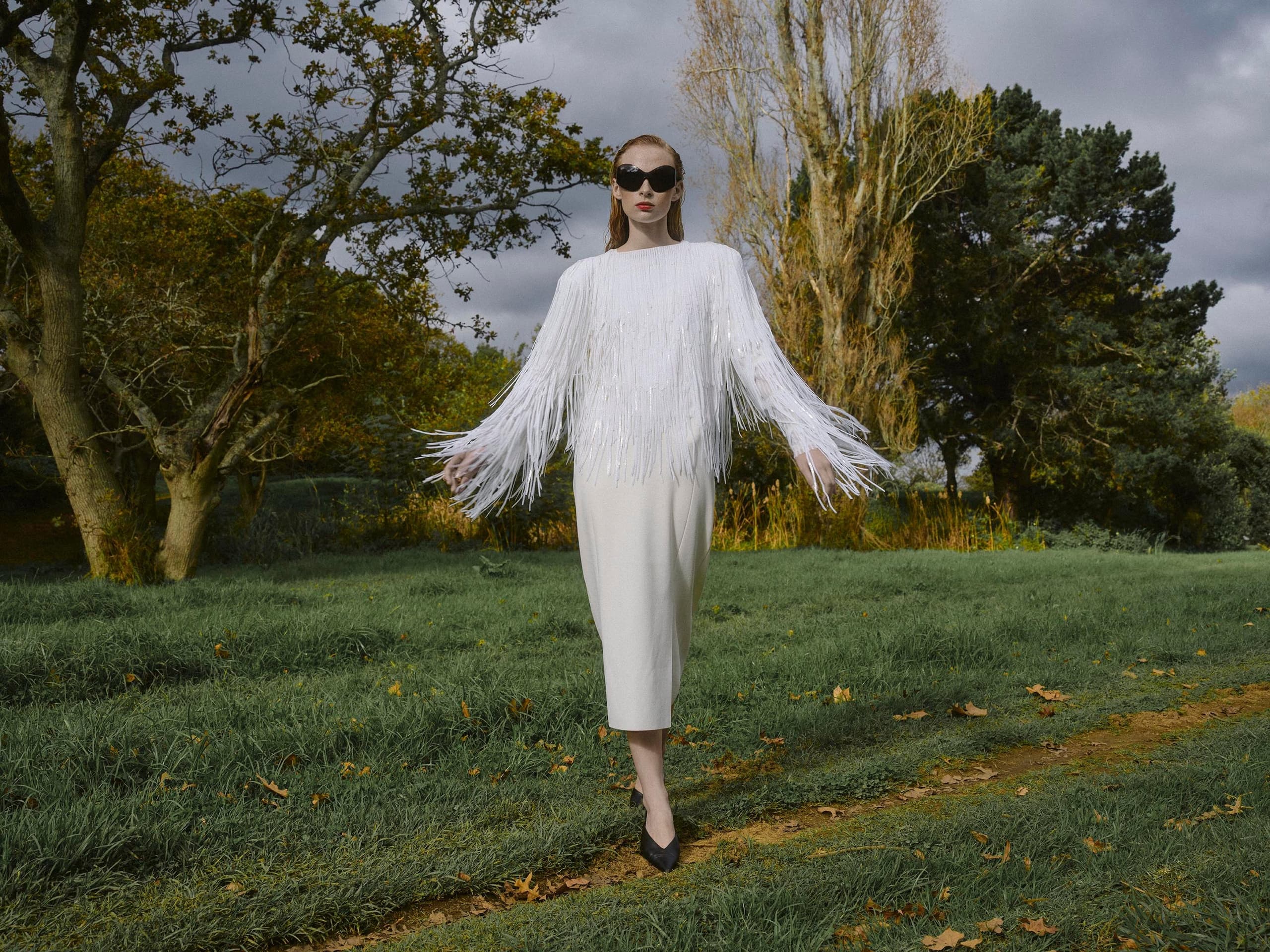 Fashionable woman in chic white fringe dress in a lush, green outdoor setting.