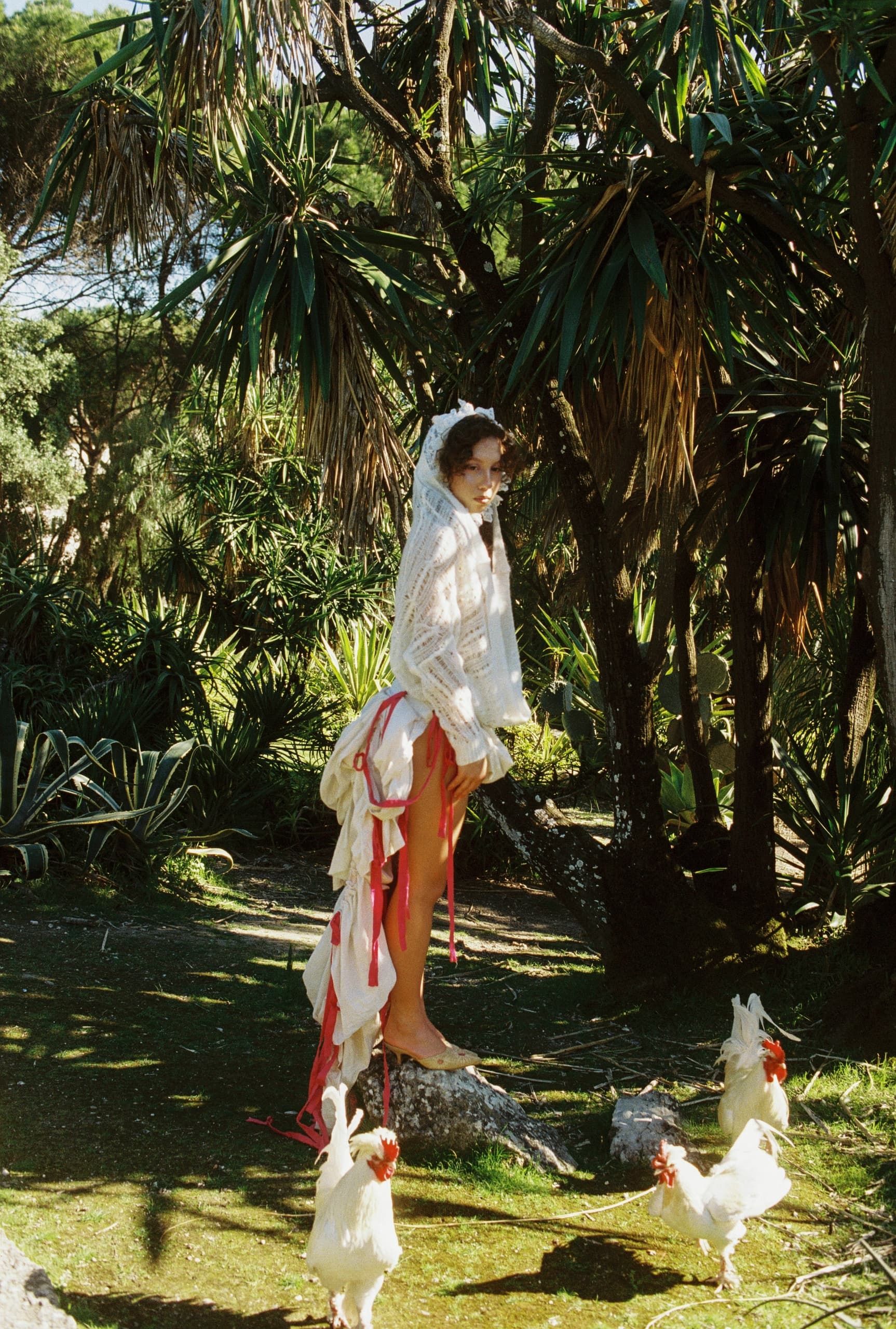 Person in avant-garde fashion with roosters in a lush tropical setting.