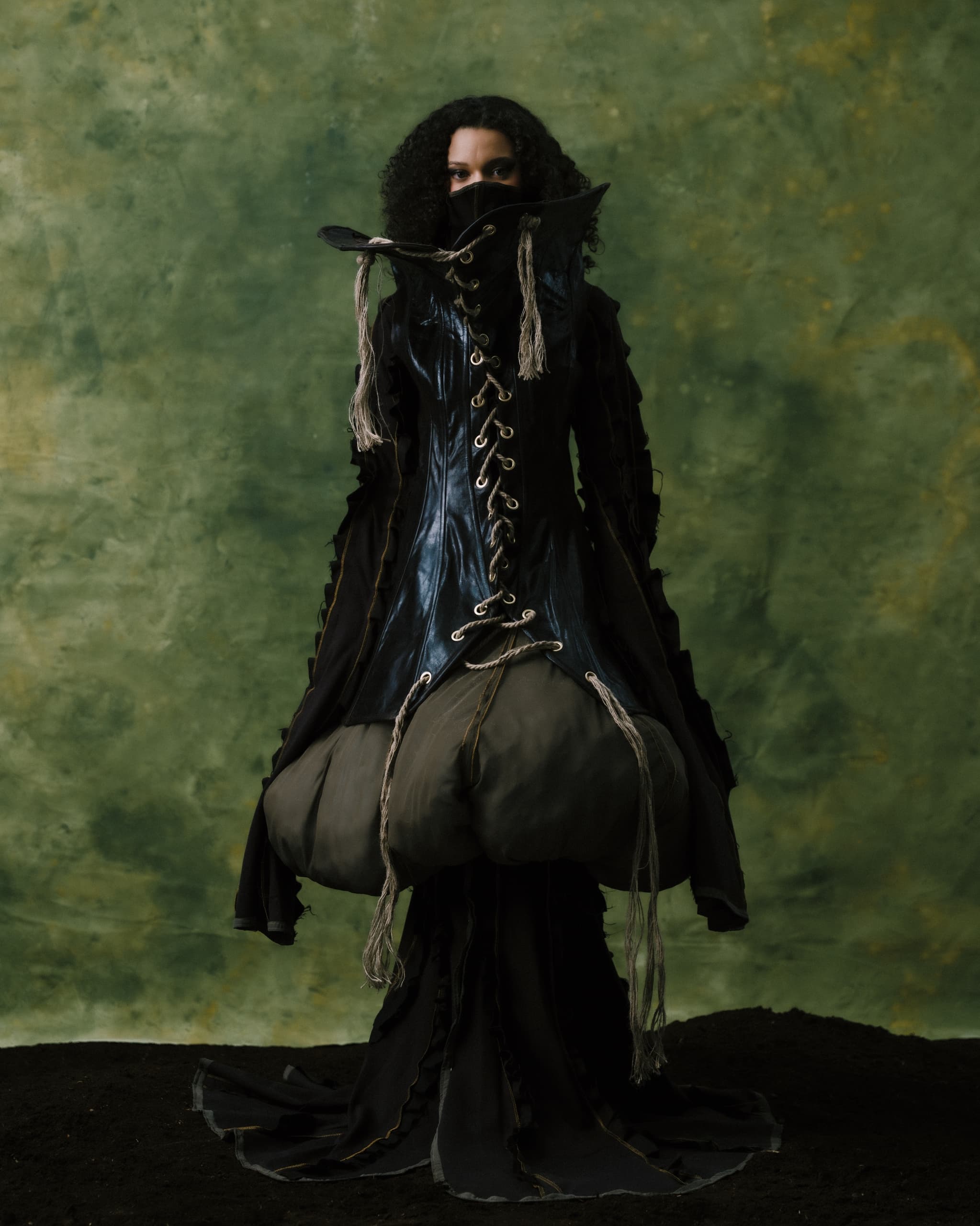 Gothic fashion portrait: person in elaborate dark, laced outfit against green backdrop.