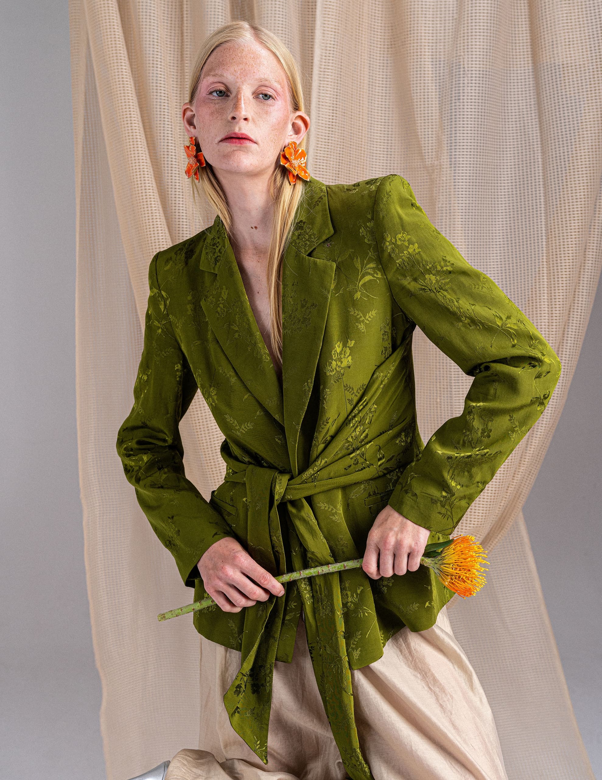 Model in green floral blazer holding a flower, against a draped curtain backdrop.