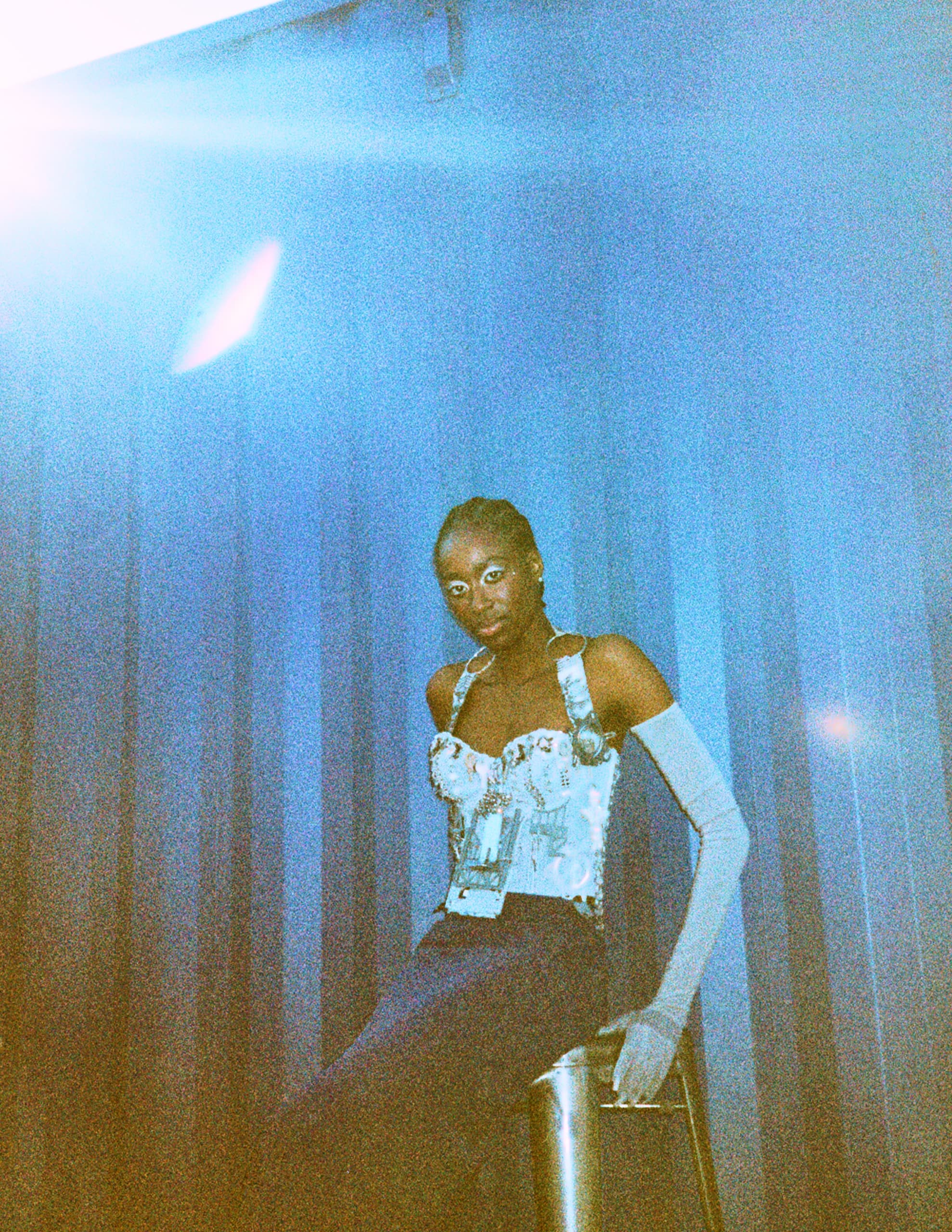 Person seated on stool in stylish outfit, blue background, dramatic lighting.