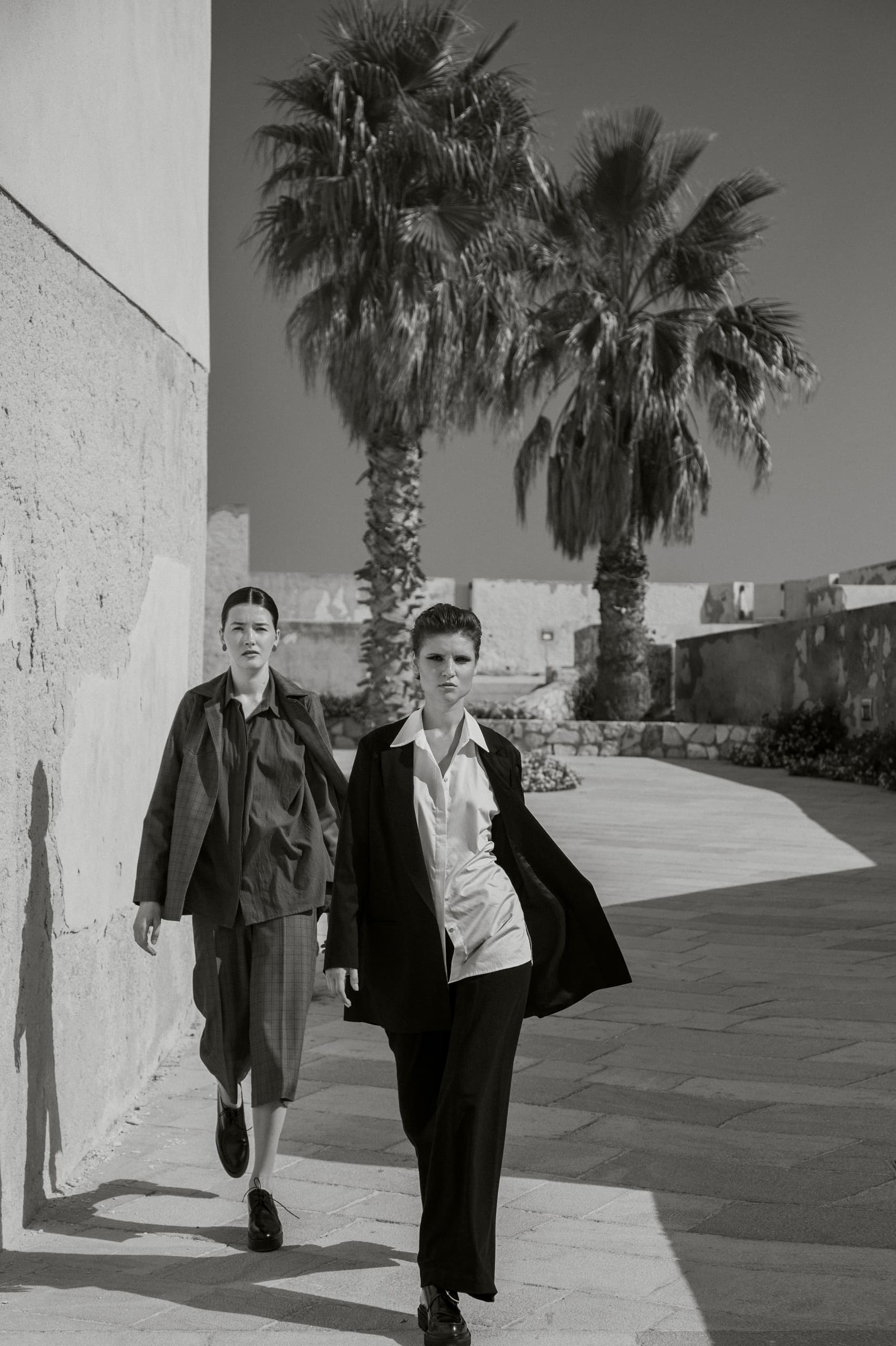 Stylish women walking under palm trees in chic outfits, black-and-white photo.