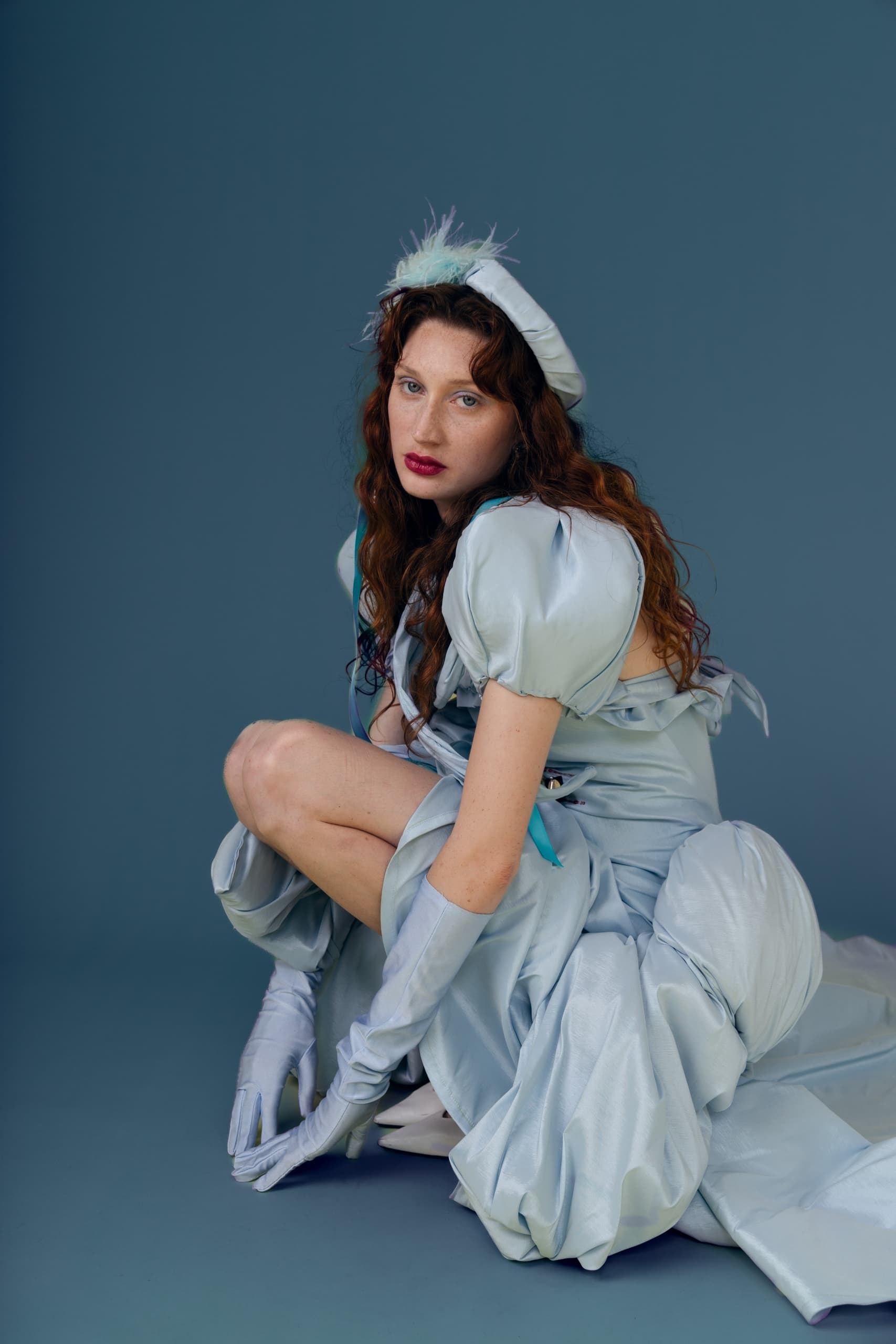 Woman in elegant blue dress and hat seated on blue background.