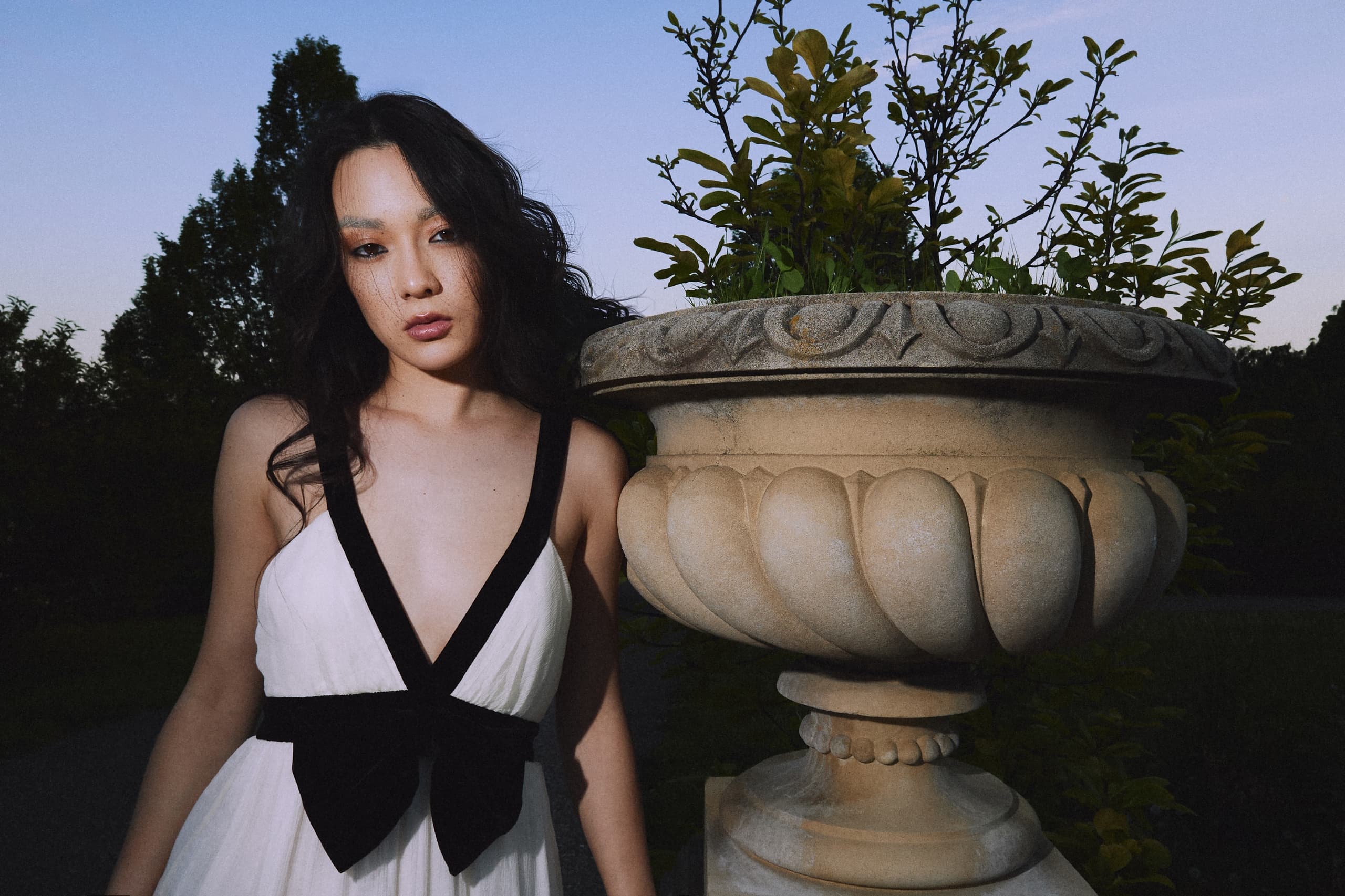 Woman in black and white dress beside ornate stone planter at dusk.
